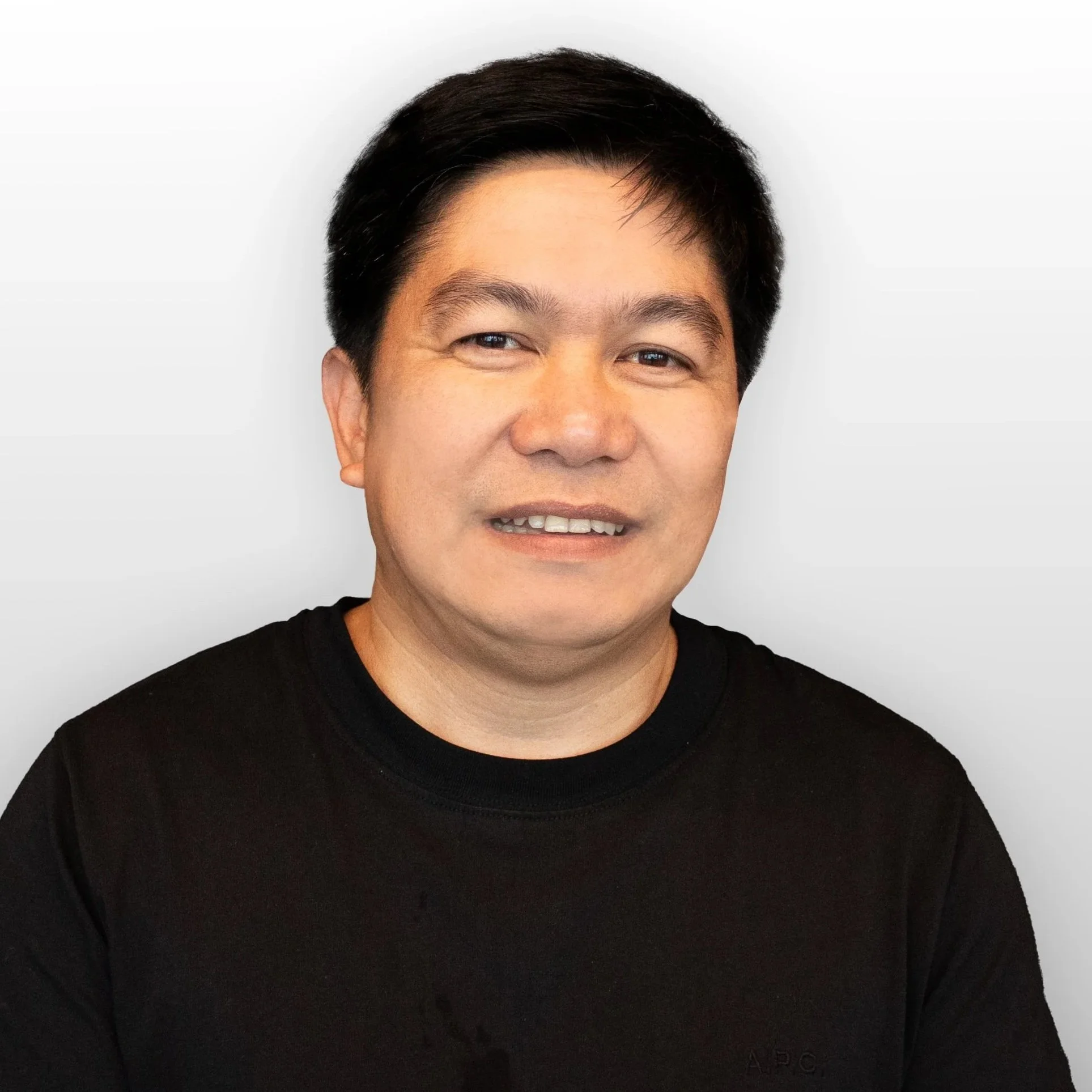 Close-up of a smiling person with short black hair wearing a black shirt, against a plain light background.
