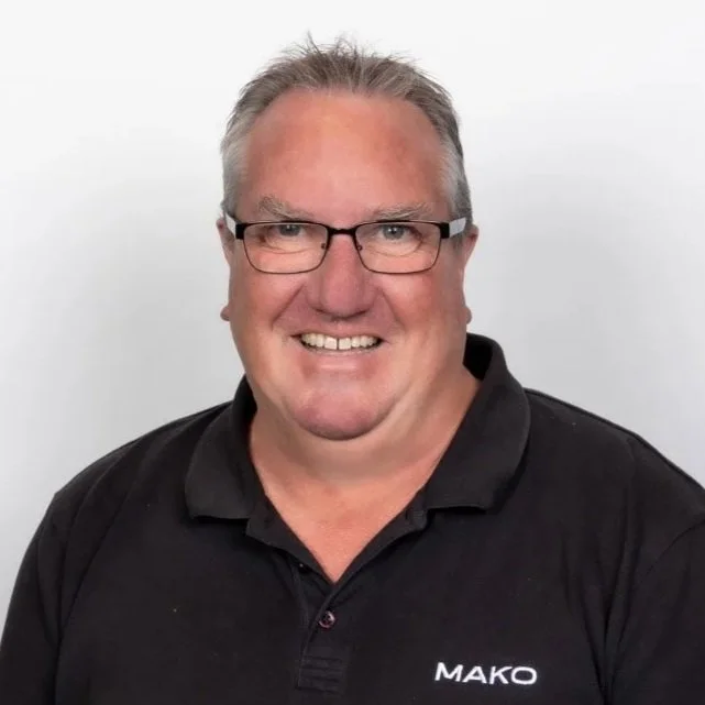A smiling middle-aged man with glasses and short gray hair, wearing a black polo shirt with the name "MAKO" embroidered on it, standing against a plain white background.