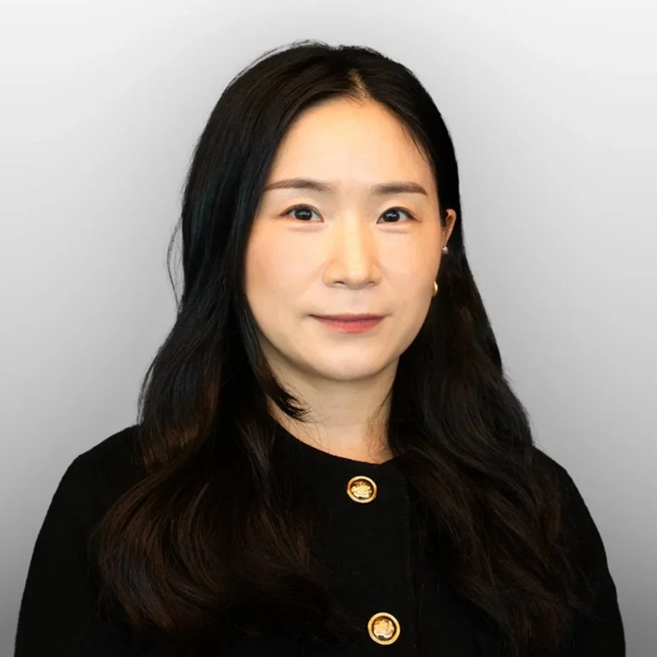 Portrait of a woman with long black hair wearing a black top with gold buttons, smiling softly against a plain gray background.