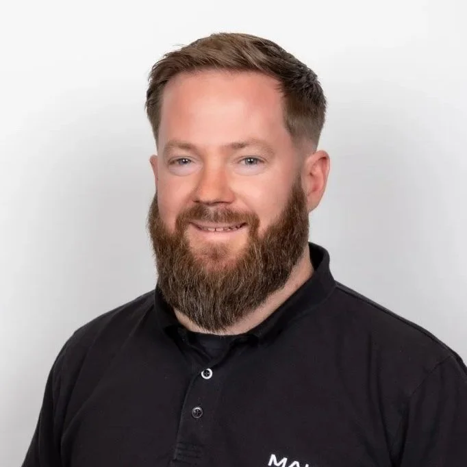 A man with a beard wearing a black collared shirt with a logo, smiling against a plain white background.