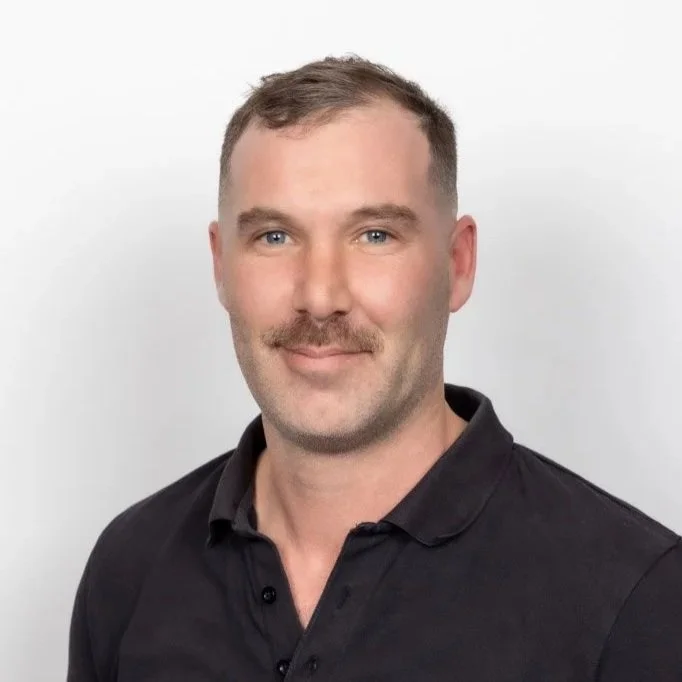 Portrait of a man with short brown hair, blue eyes, light complexion, wearing a black polo shirt, against a plain white background.