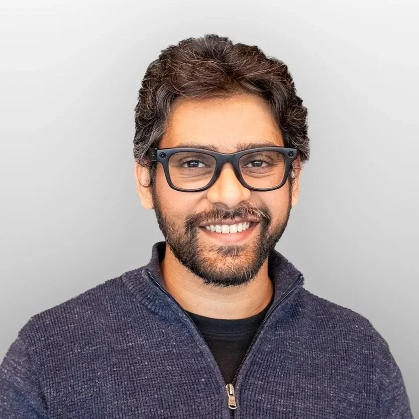 A man with dark hair, beard, and glasses smiling at the camera against a plain light gray background.