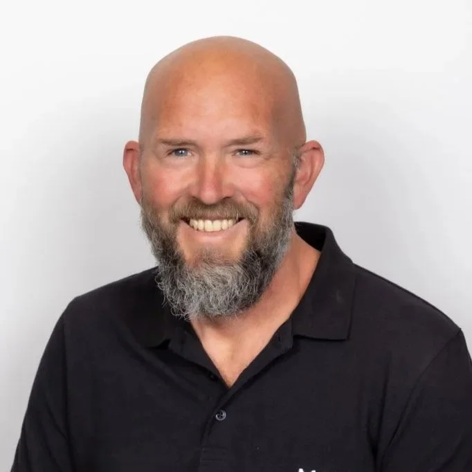 Smiling bald man with a beard wearing a black collared shirt against a white background.