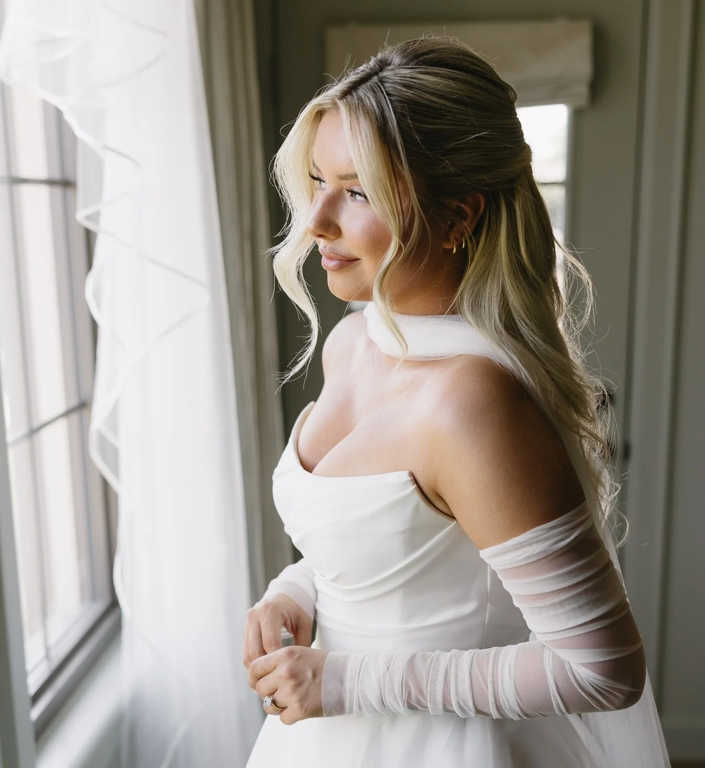 A woman in a wedding dress standing near a window, gazing outside with a gentle smile.