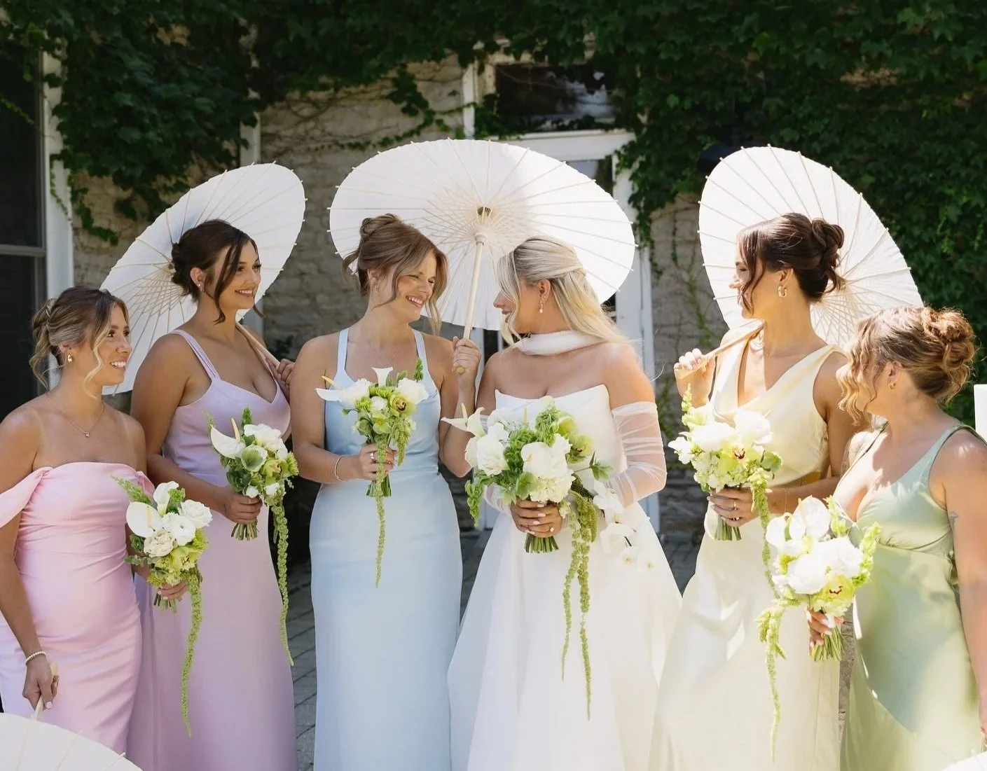 Group of women in pastel dresses holding bouquets and white parasols, standing outdoors against a stone building and greenery.