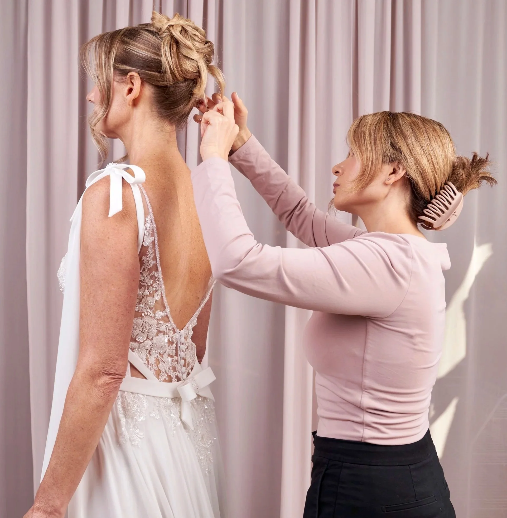A woman trying on a wedding dress while a stylist adjusts her hair in front of a light-colored curtain.