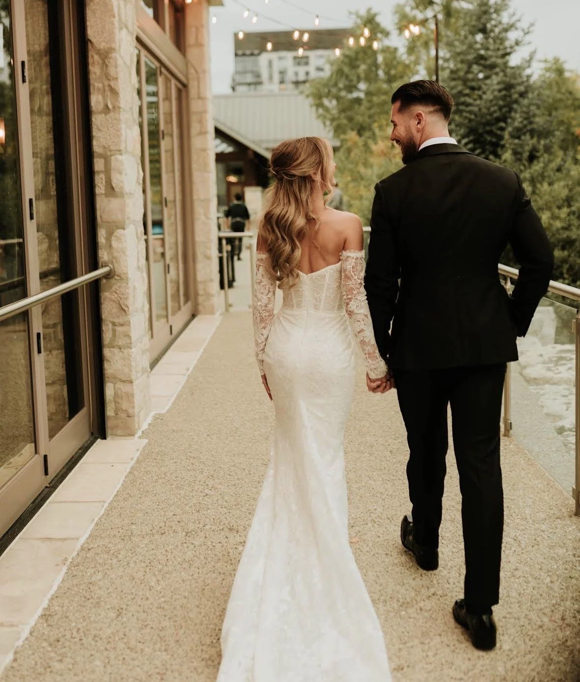 A bride and groom walking hand in hand on a terrace during their wedding celebration, with trees and string lights overhead.