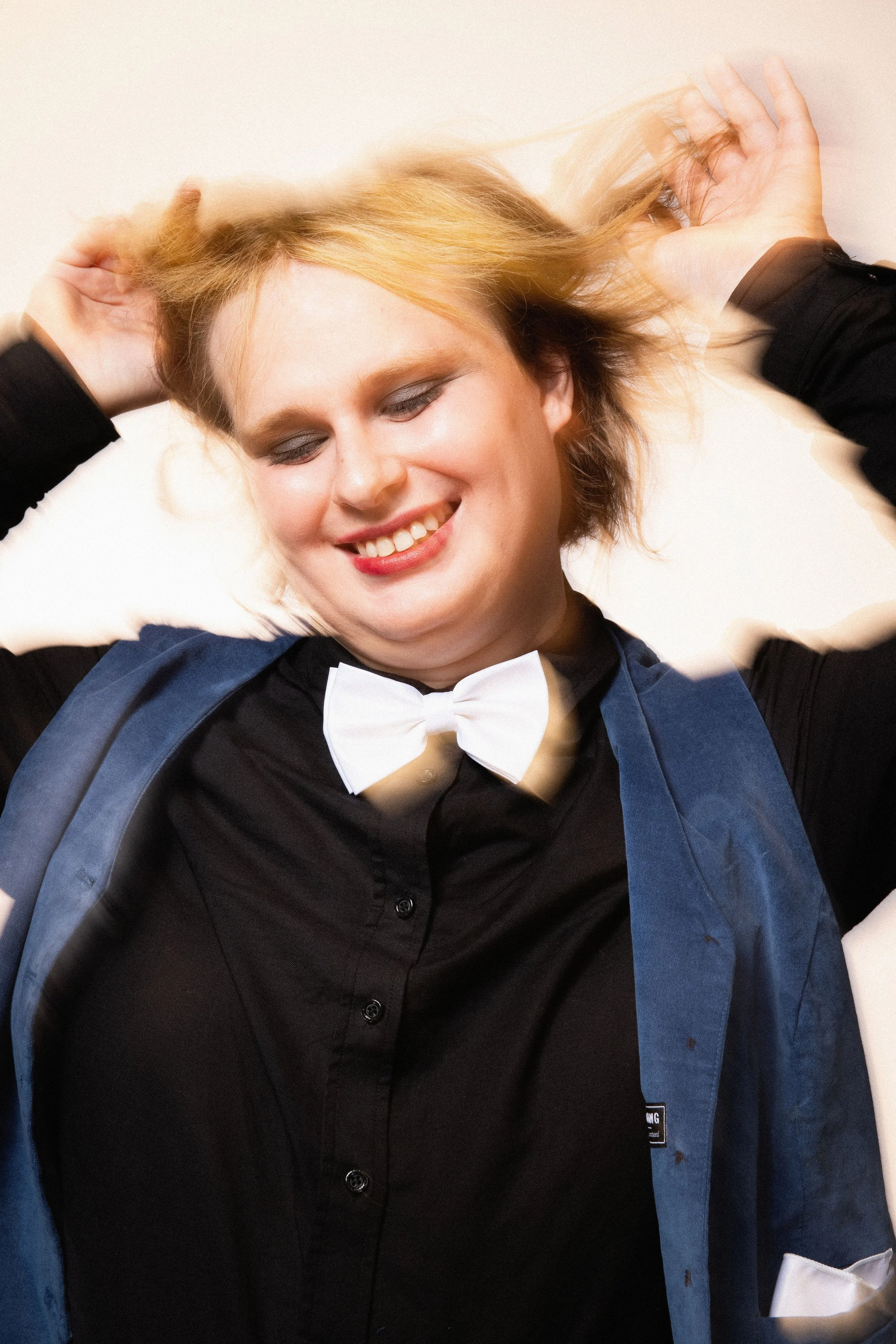 A young woman with blonde hair holding her hair back, smiling, dressed in a tuxedo with a white bow tie and a blue jacket, against a plain background.