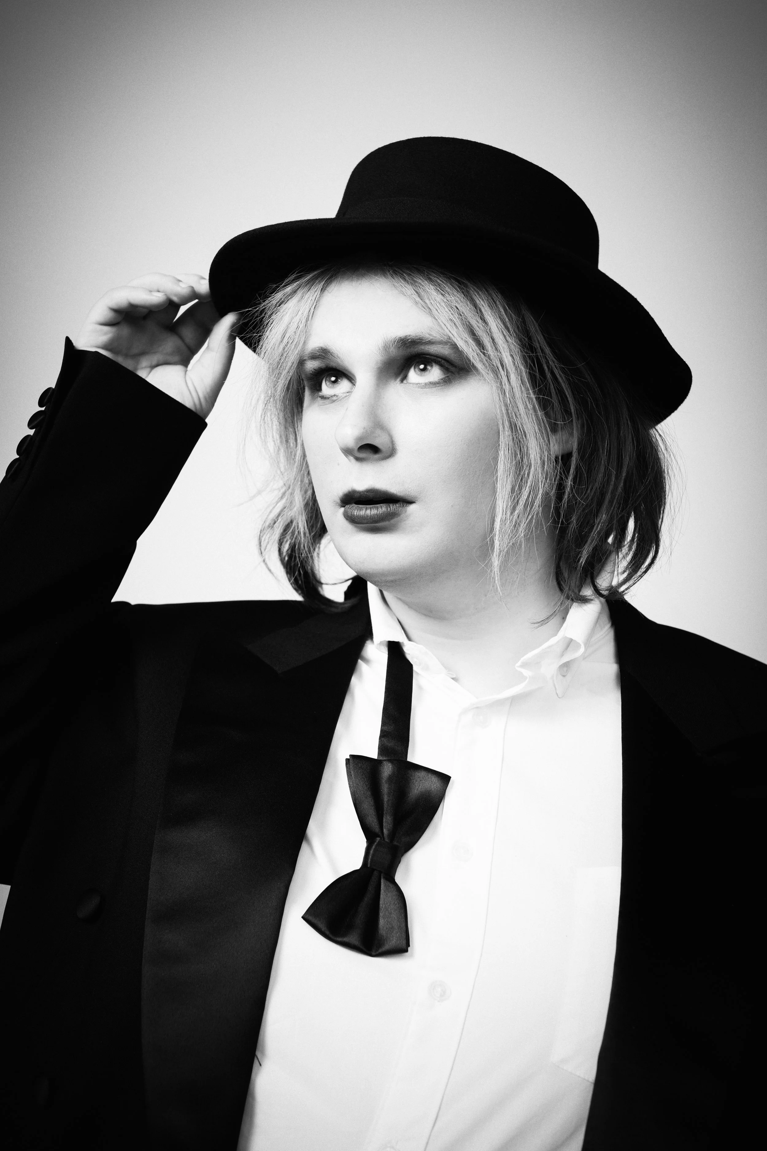 Black and white portrait of a woman adjusting a wide-brimmed hat. She has shoulder-length hair, is wearing a tuxedo with a white shirt, and a black bow tie. The background is plain and gradient.