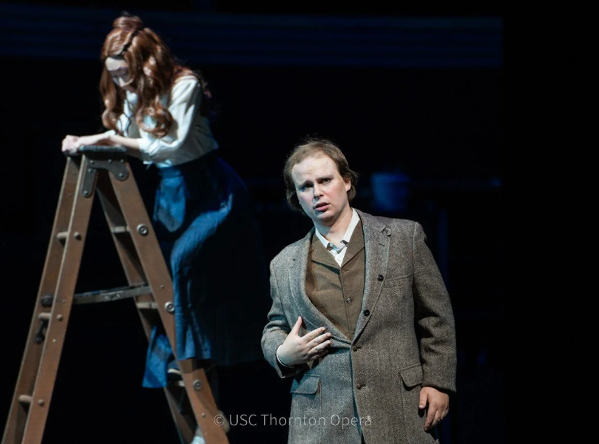 A stage scene with two actors; one woman on a ladder looking down, and a man standing nearby with a hand on his chest, appearing emotional.