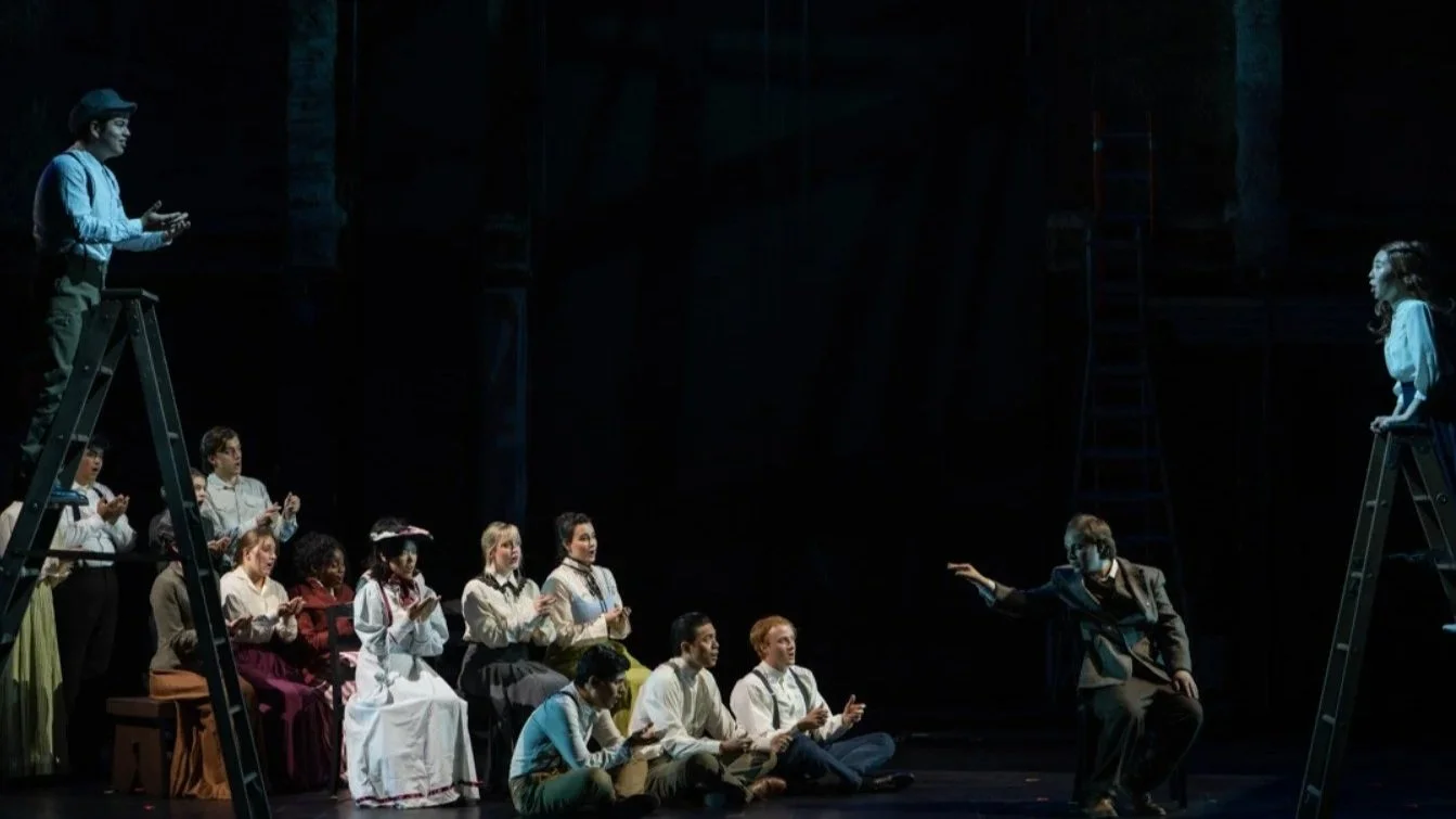 Theater stage scene depicting a diverse group of actors, some standing on ladders, others seated, with a director or conductor kneeling and gesturing towards a woman on the right ladder. The stage is dark with minimal lighting focused on the performers.