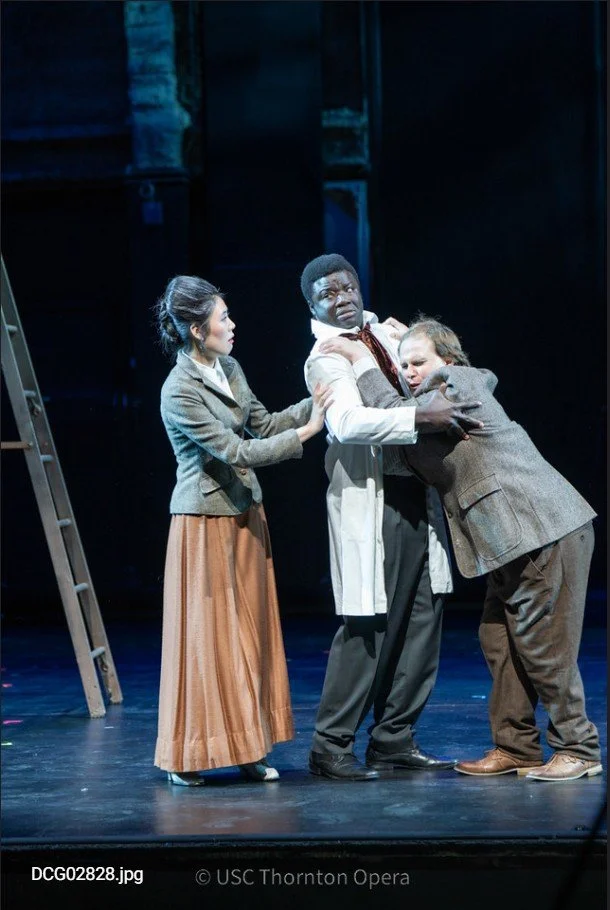 Three performers on stage, engaged in a dramatic scene. The woman on the left is holding the arm of the man in the center, who appears distressed. A third man is hugging the center man tightly. The background is dark with stage lighting, and there is a ladder on the left side.