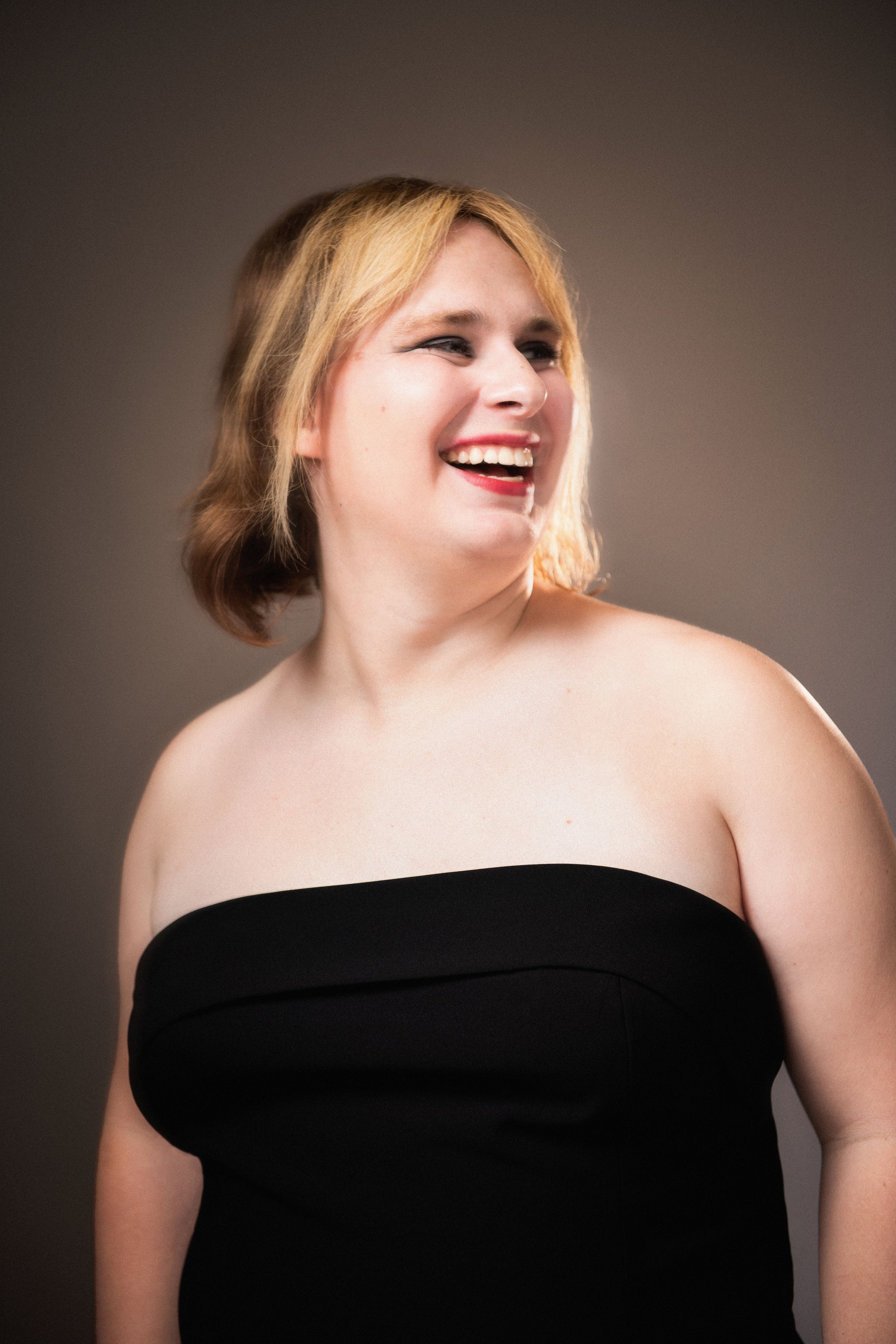 A woman smiling, wearing a strapless black top, with short blonde hair against a dark background.