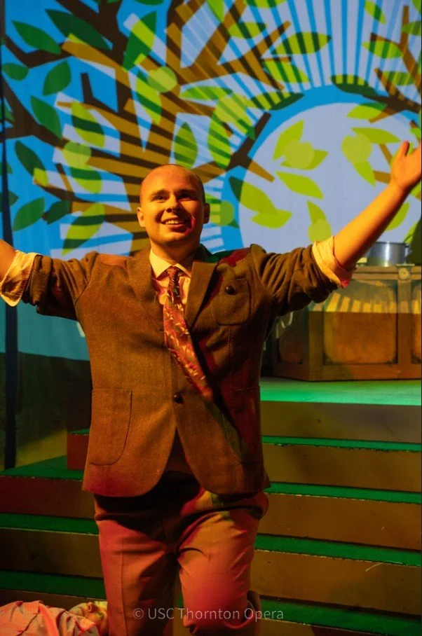 A man in a brown suit with a colorful tie, smiling with arms raised, standing in front of a stage with a vibrant, colorful projection of a tree with green leaves and brown branches in the background.