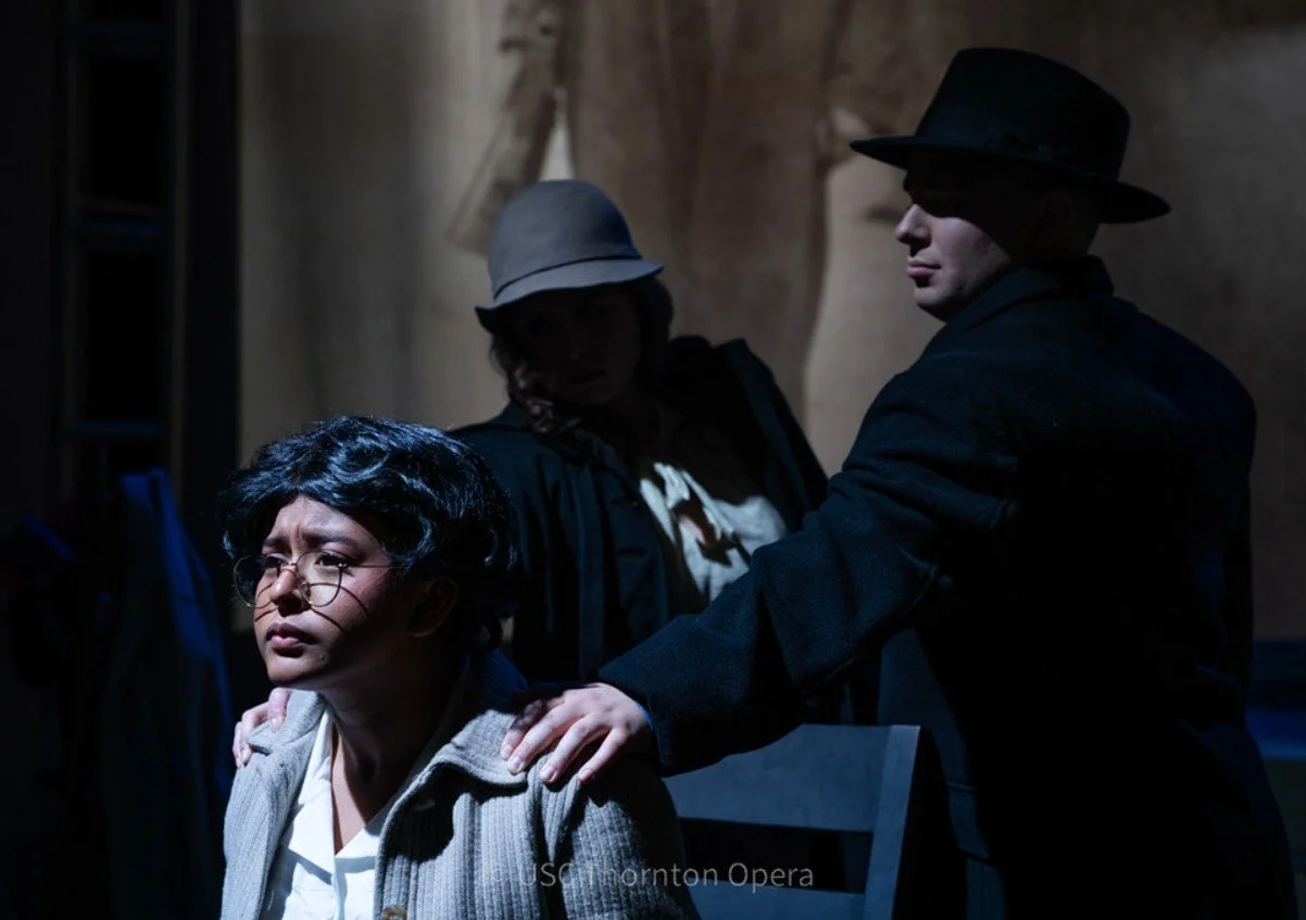 Three people in vintage clothing seated and standing in a dimly lit room. The person in the foreground has black, curly hair, glasses, and wears a light-colored sweater. A person in the middle wears a hat and dark clothing, holding the shoulder of the person in front. The third person at the back wears a cap and a scarf, looking at the others.