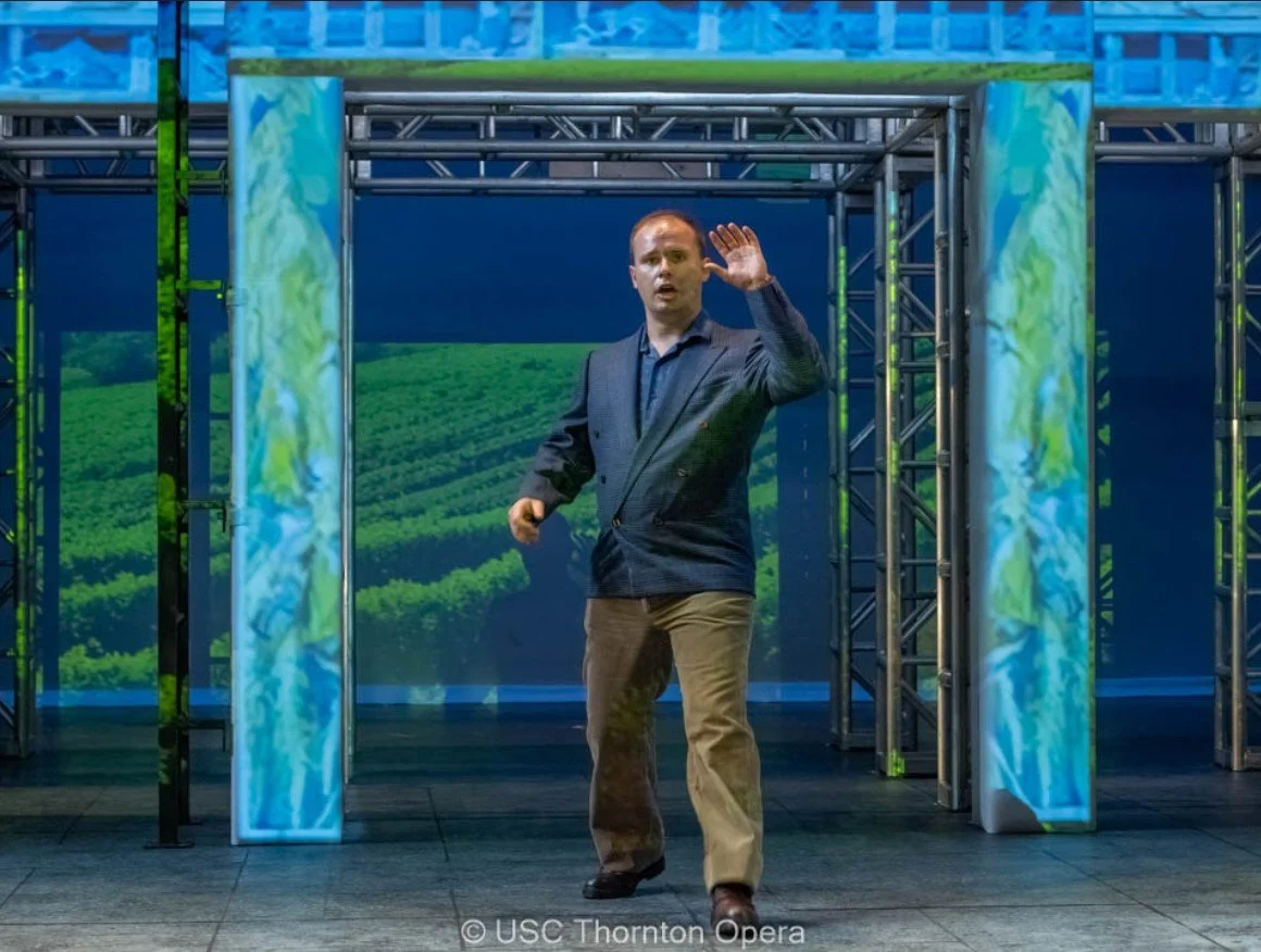 A man in a dark blazer and khaki pants standing on stage with a blue backdrop and green landscape projection, gesturing with his right hand raised and open.