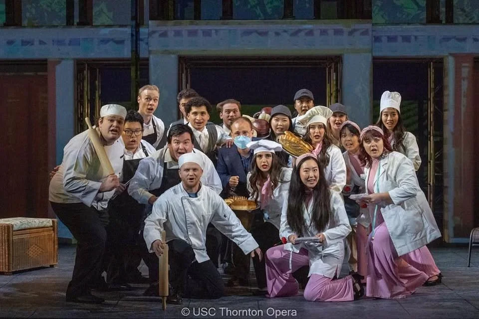 A group of actors on stage in a theatrical production, dressed as chefs, nurses, and other characters, with some holding props and costumes.