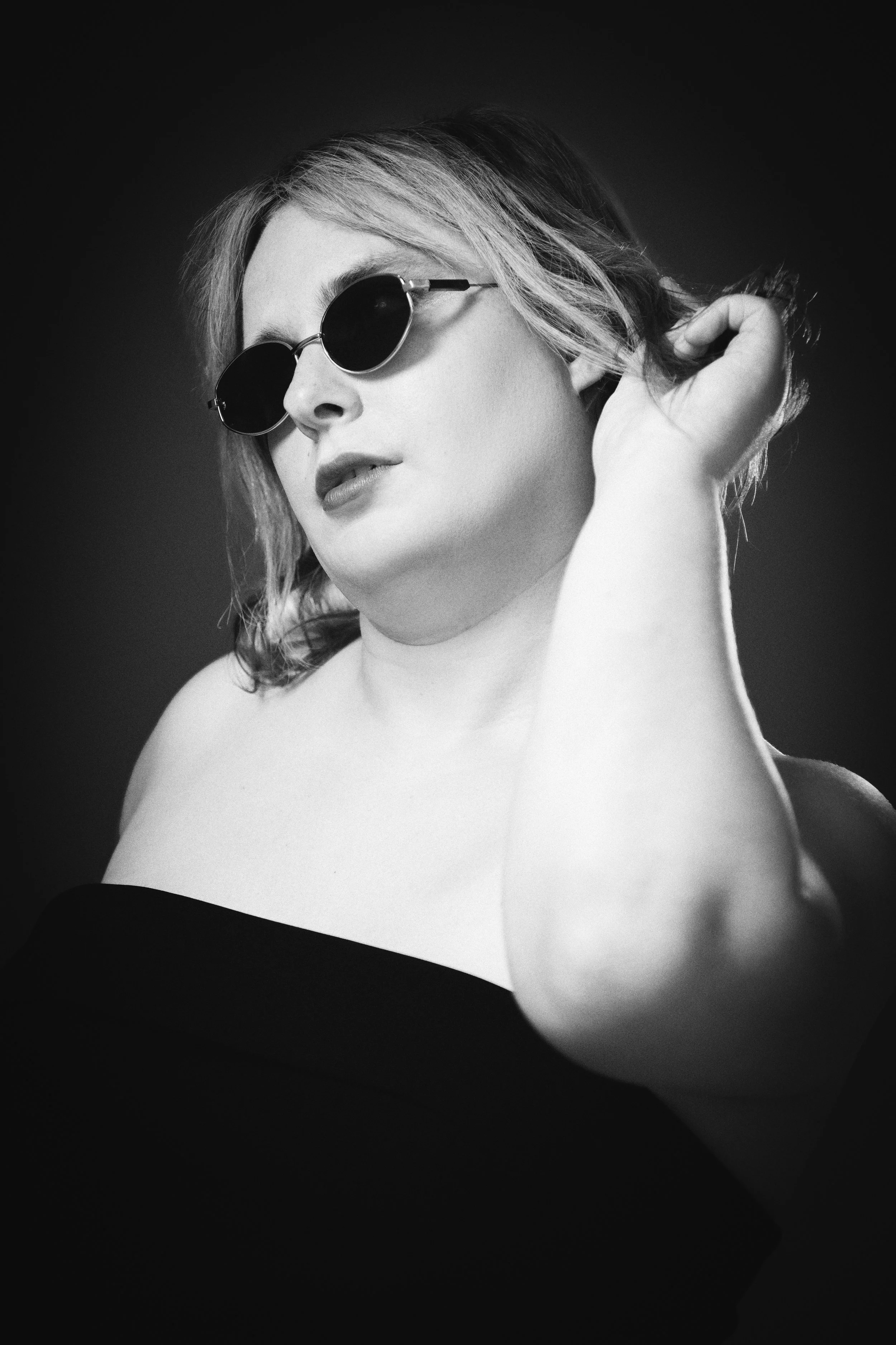 A black and white photo of a woman wearing round sunglasses, touching her hair, with her bare shoulder and upper chest visible, against a dark background.