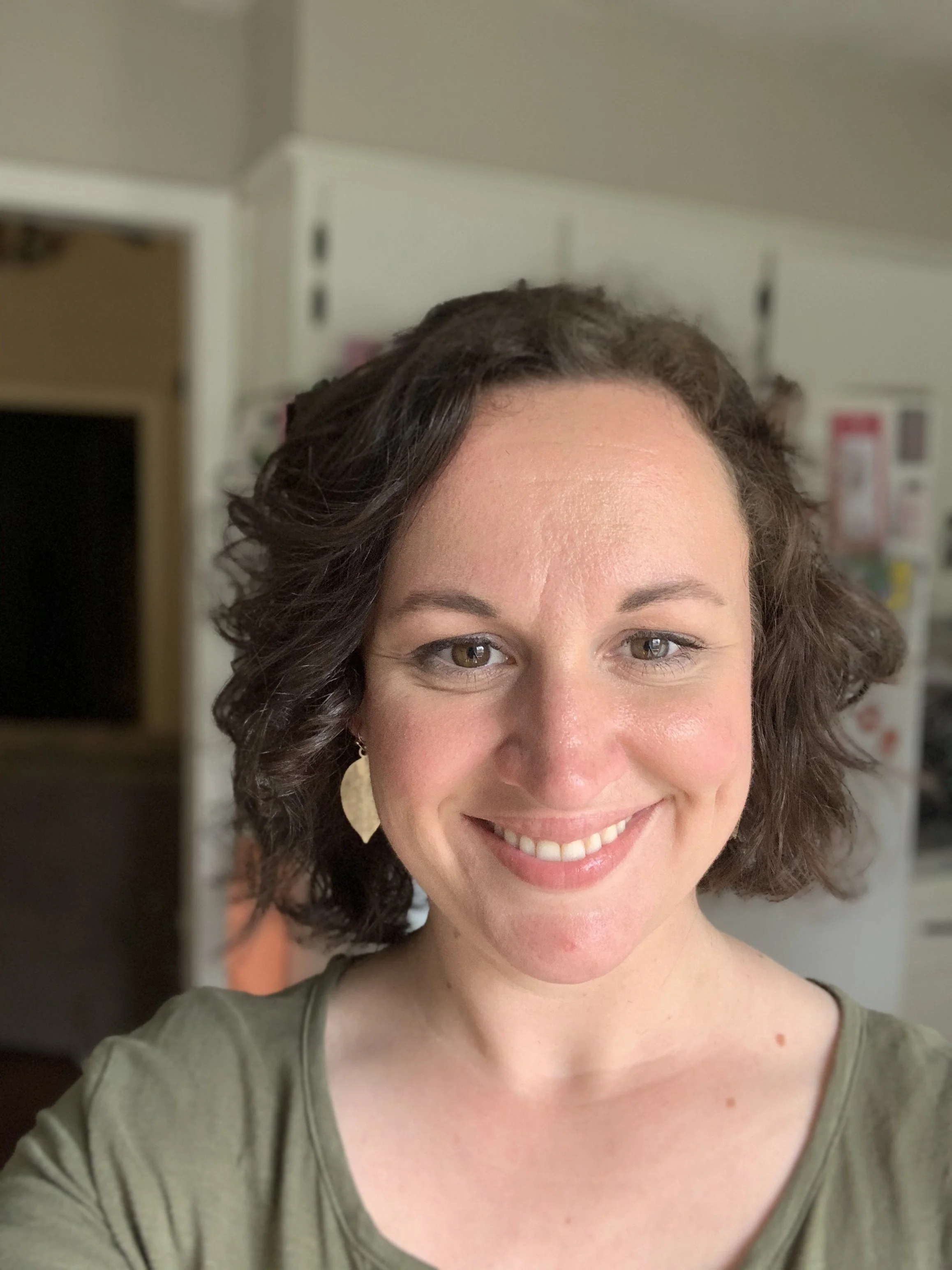 Close-up of a smiling woman with curly brown hair, wearing a green top, in a room with white cabinets in the background.