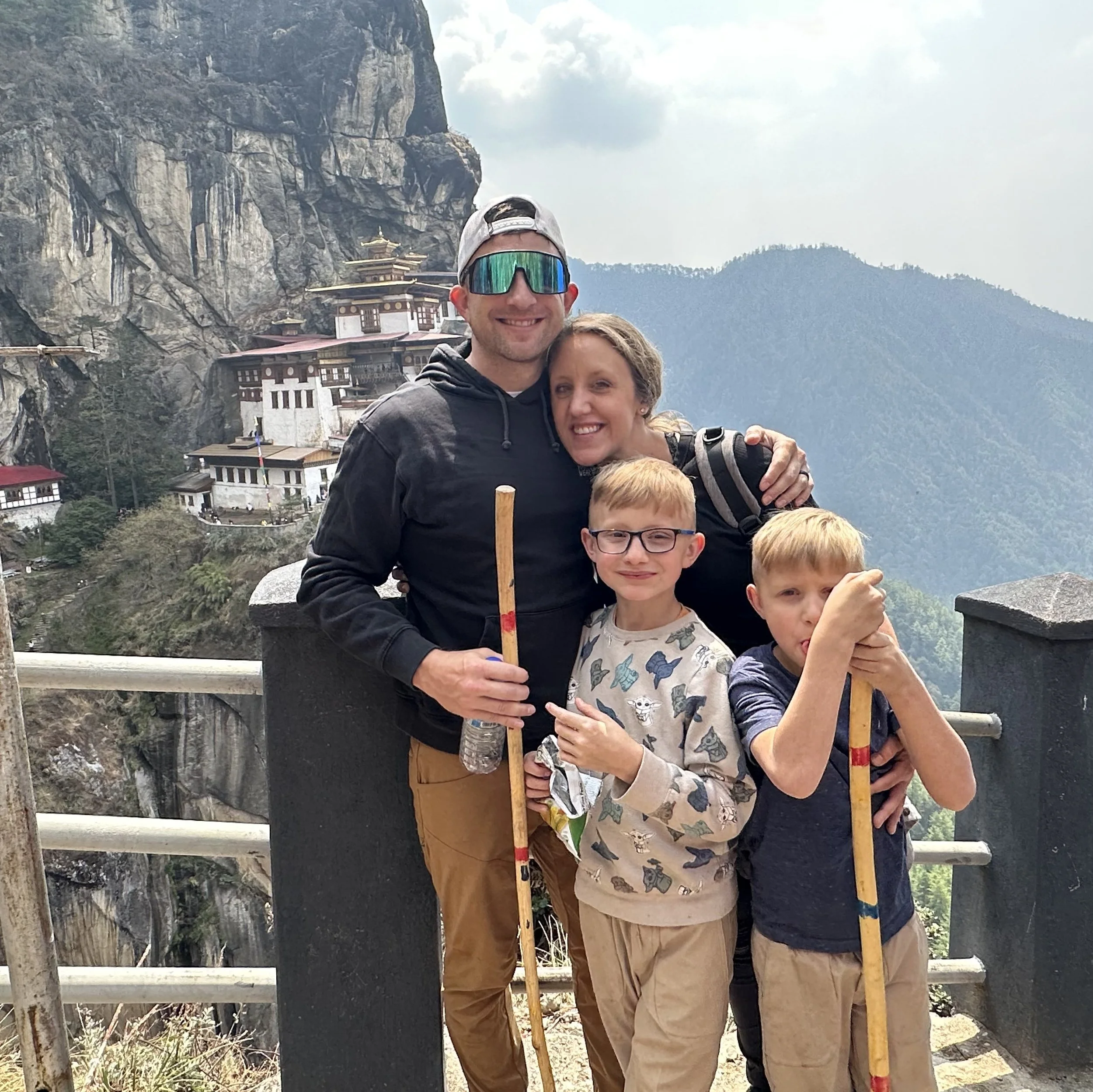 A group of people, including children and adults, standing together on a mountain ledge, with traditional Himalayan-style buildings and cliffs in the background.