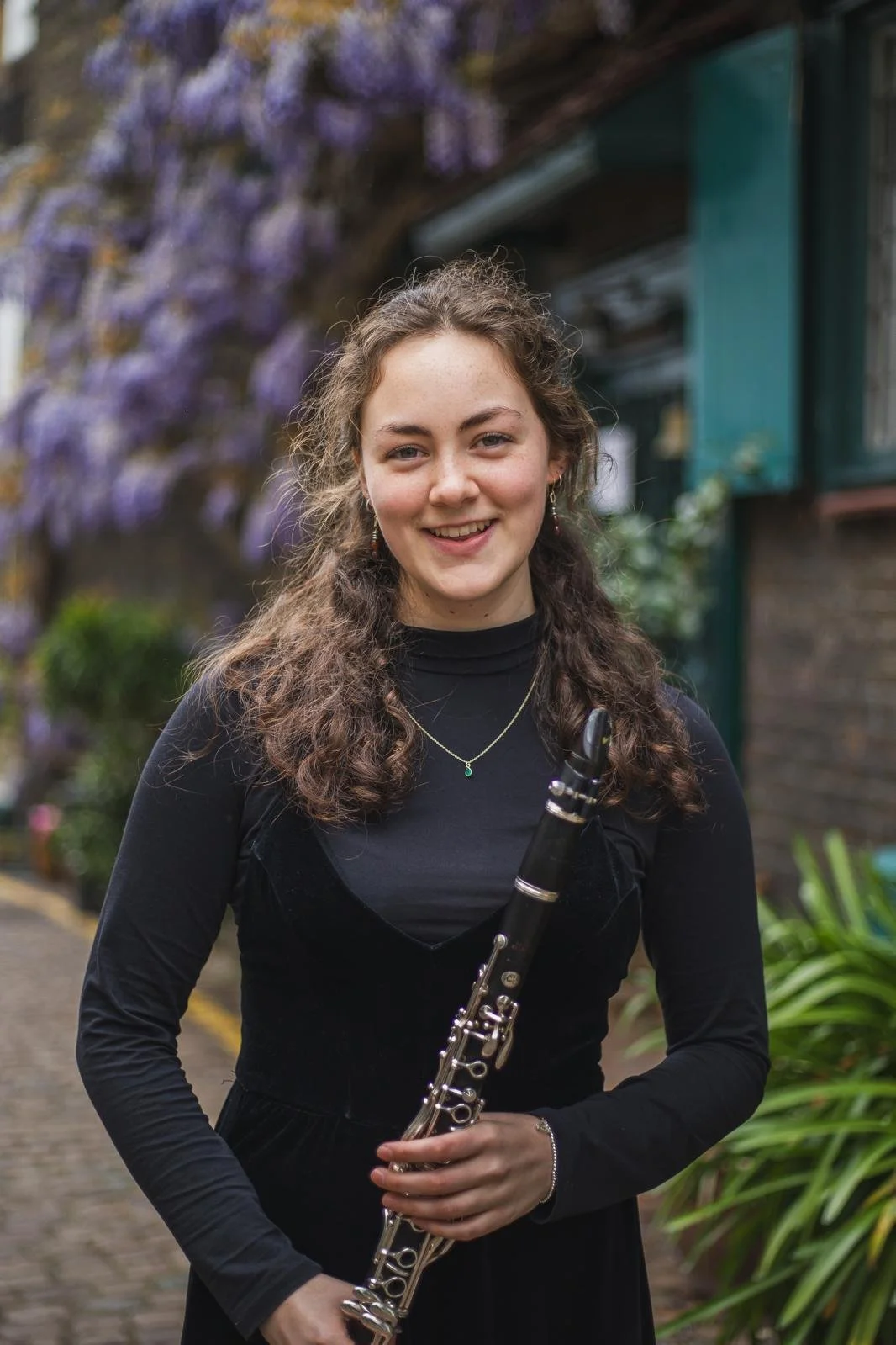 Young woman with curly hair holding a clarinet, standing outdoors with purple flowering tree and greenery in the background.