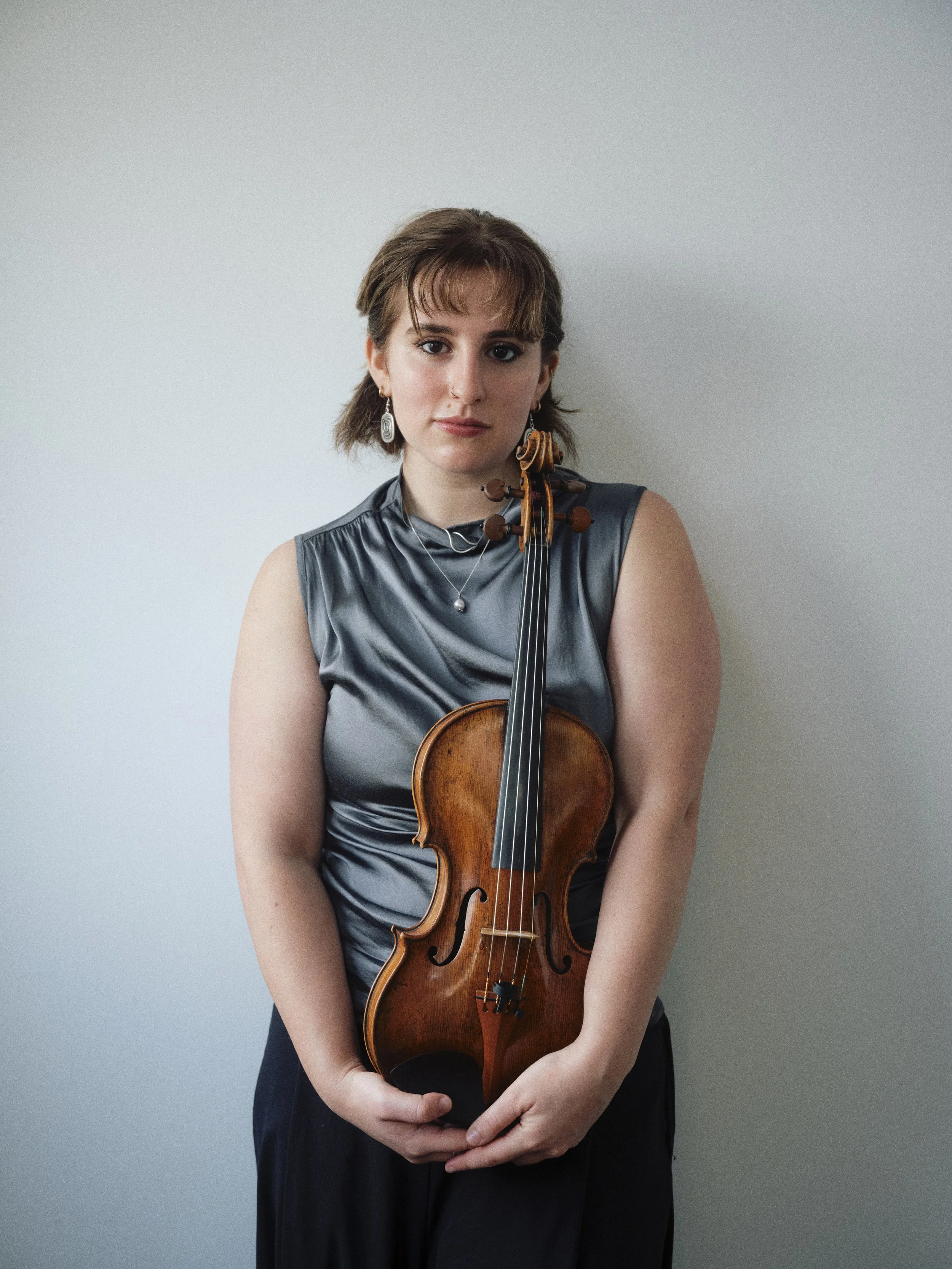 A woman with short brown hair and earrings holds a violin in front of a plain gray wall.