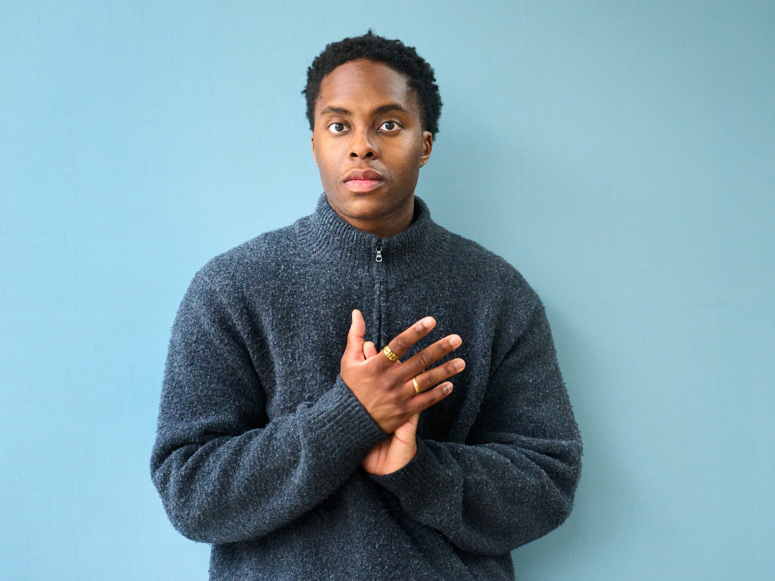 Portrait of a young man with dark curly hair, wearing a gray zip-up sweater, standing against a light blue wall, with his left hand resting on his chest and his right hand holding his left wrist.