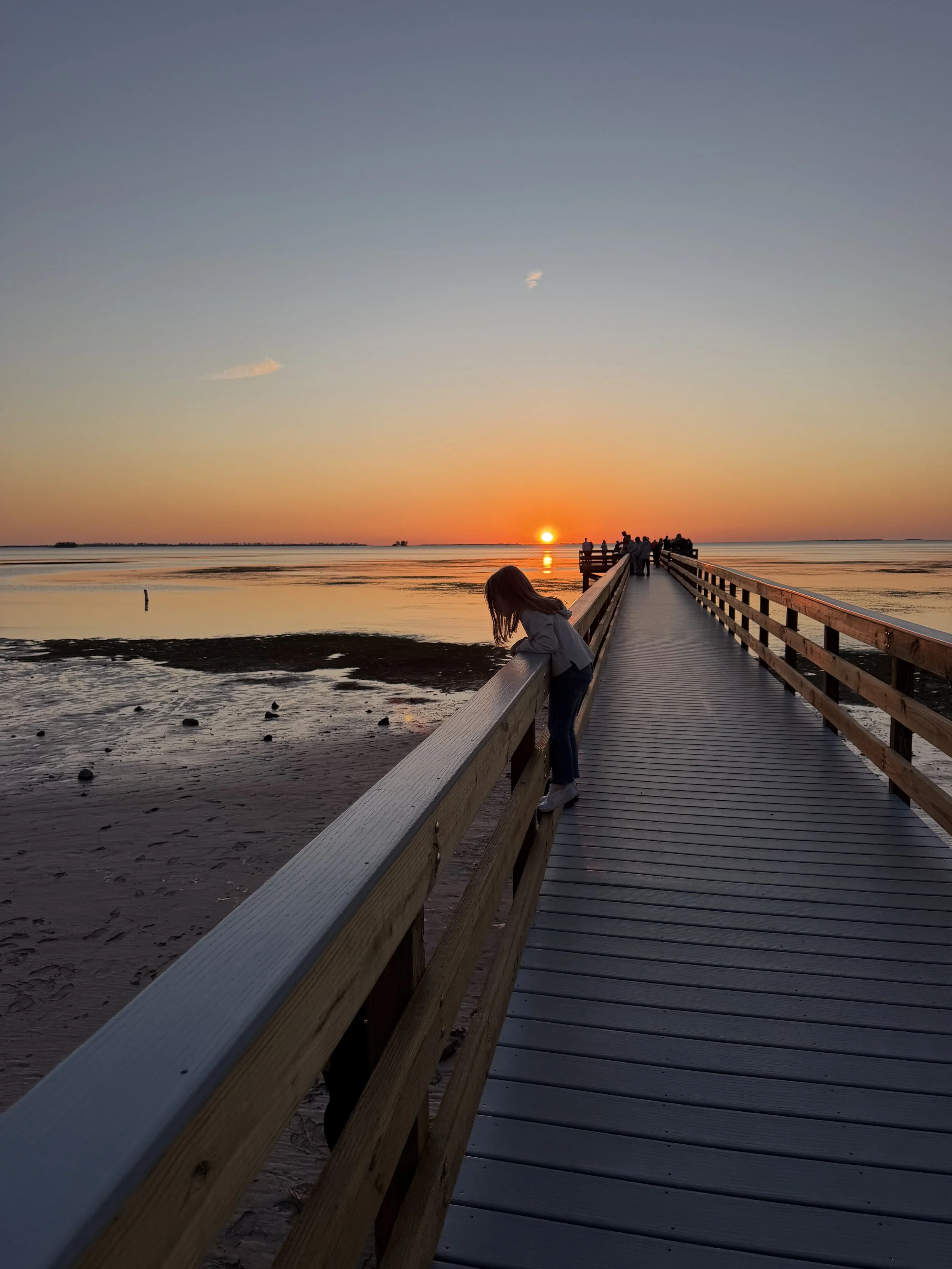 A wooden pier extending over a body of water at sunset. People are gathered at the end of the pier, and a young girl leans on the railing looking down. The sky is a gradient of warm colors with the sun near the horizon.