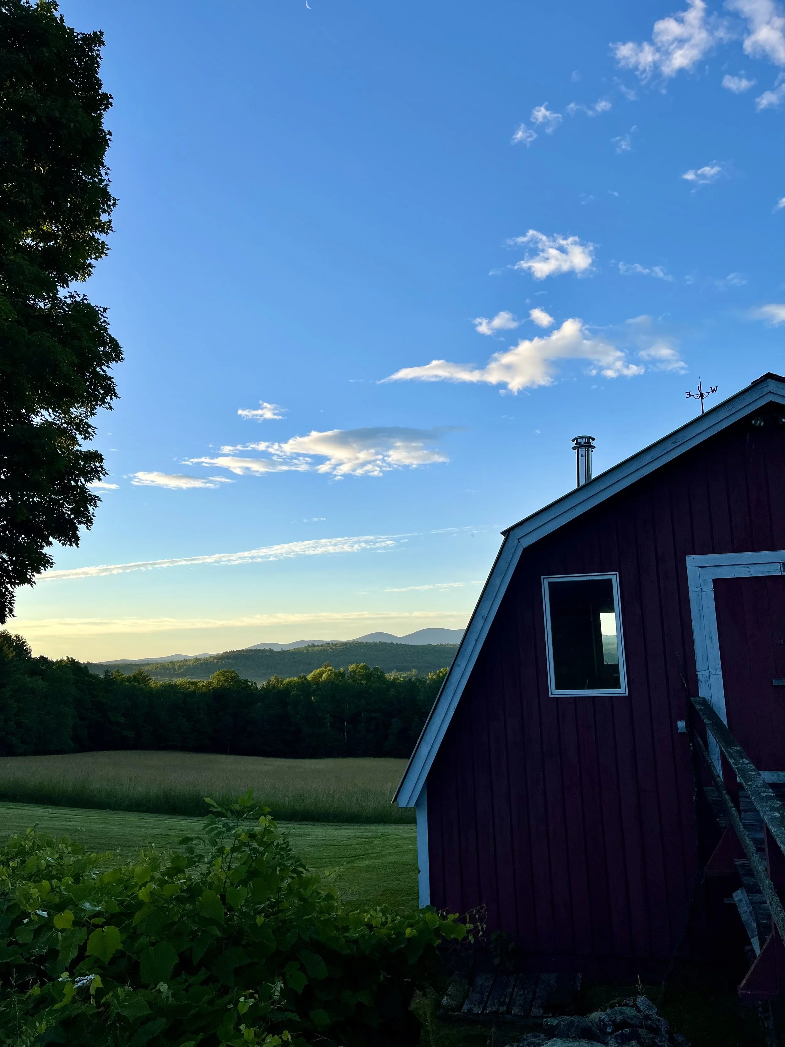 Sunset sky with a few clouds over a red barn with a ladder leaning against it, surrounded by green trees and fields, mountains in the distance.