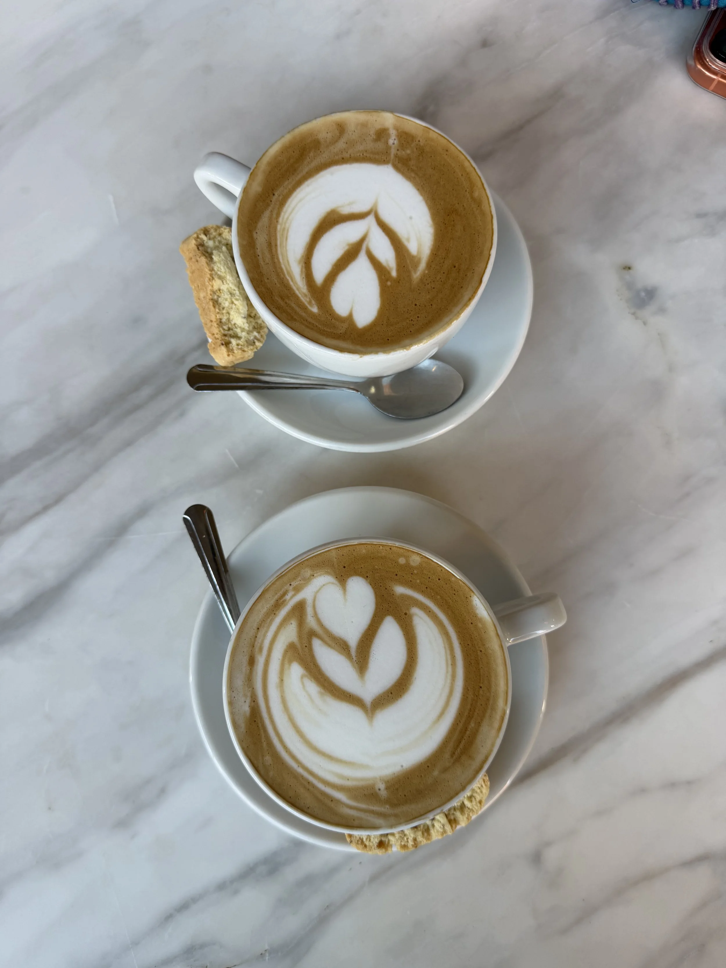Two cups of coffee with latte art on a white marble table, each with a biscotti cookie on the saucer and a spoon. The latte art features symmetrical heart and leaf patterns.