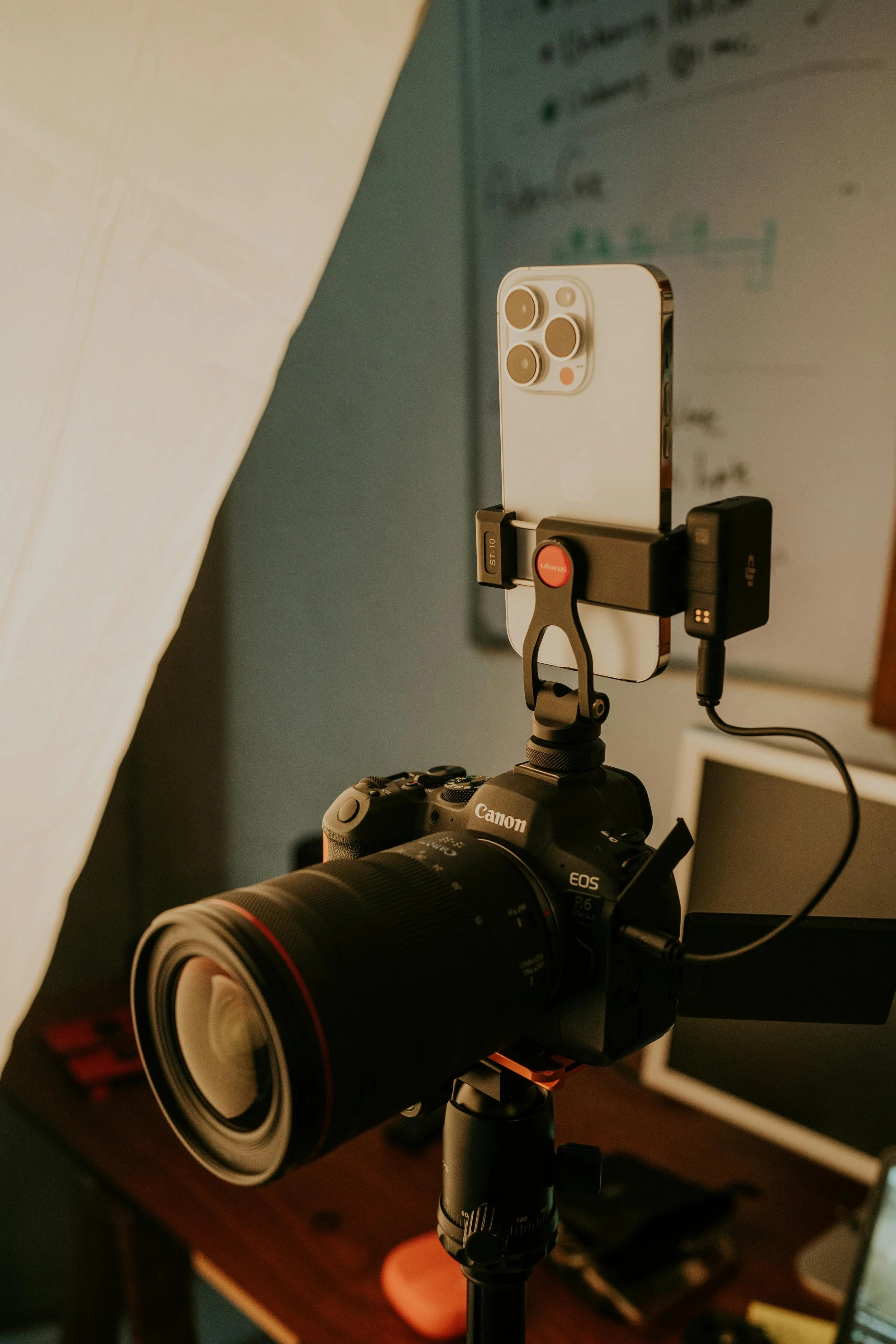 A professional camera mounted on a tripod with a smartphone attached, used for photography or videography, in a room with a computer monitor in the background.