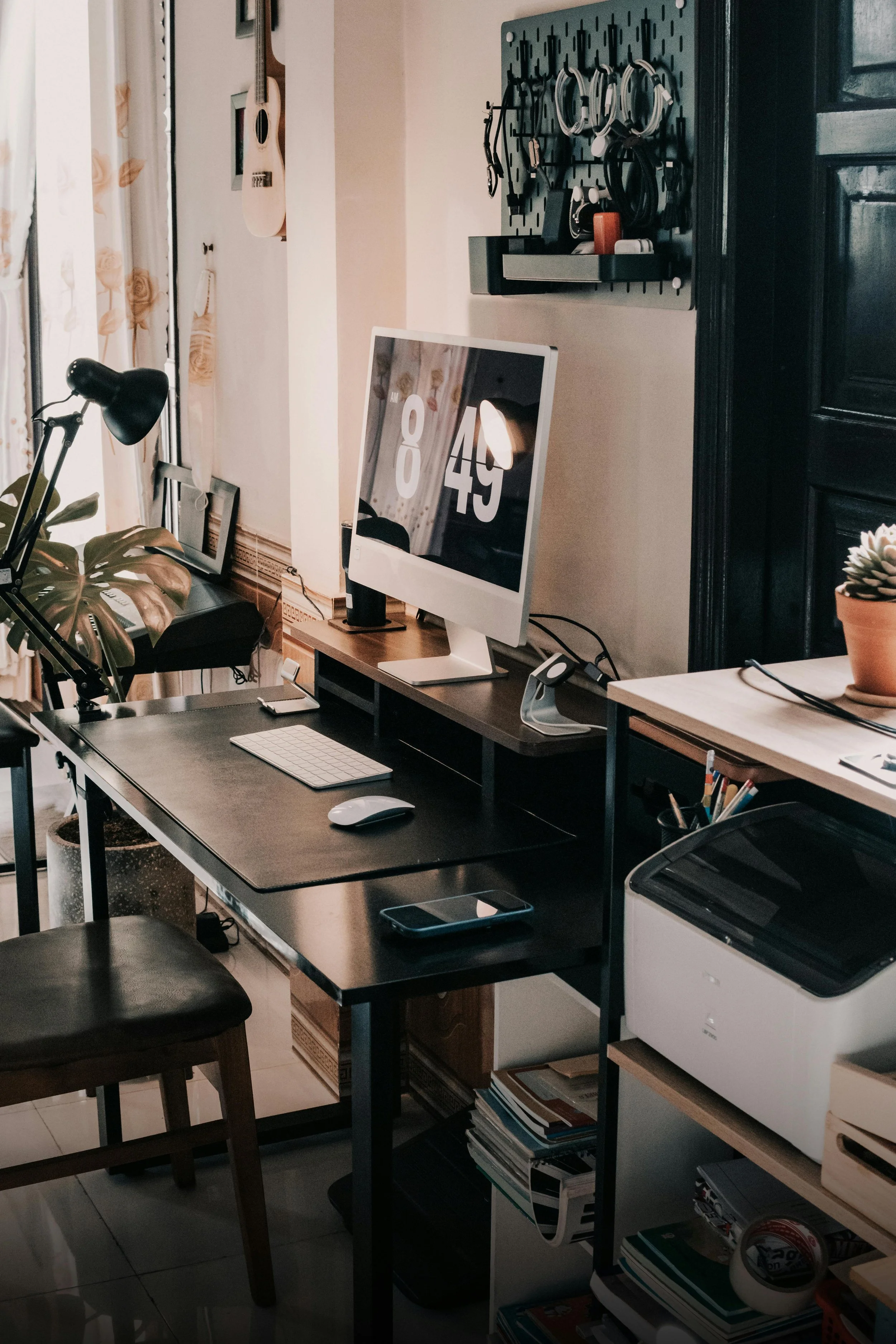 A home office with a computer monitor displaying time, a desk with a keyboard, mouse, and a smartphone. There are various shelves and hooks with accessories, cords, and storage containers. Natural light enters through a window with floral curtains.