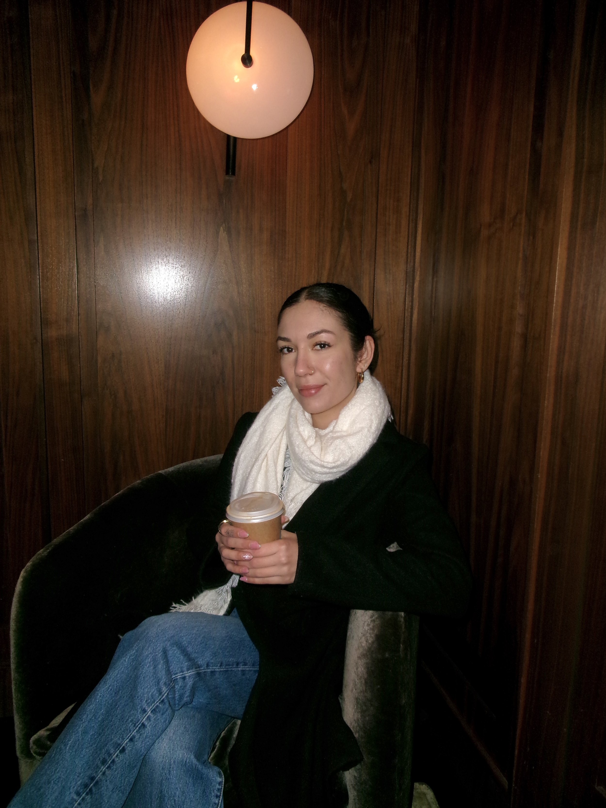 A woman with braided dark hair, wearing a white scarf, black coat, and gold hoop earrings, sitting in a wooden-paneled room, holding a paper coffee cup, with a warm smile.