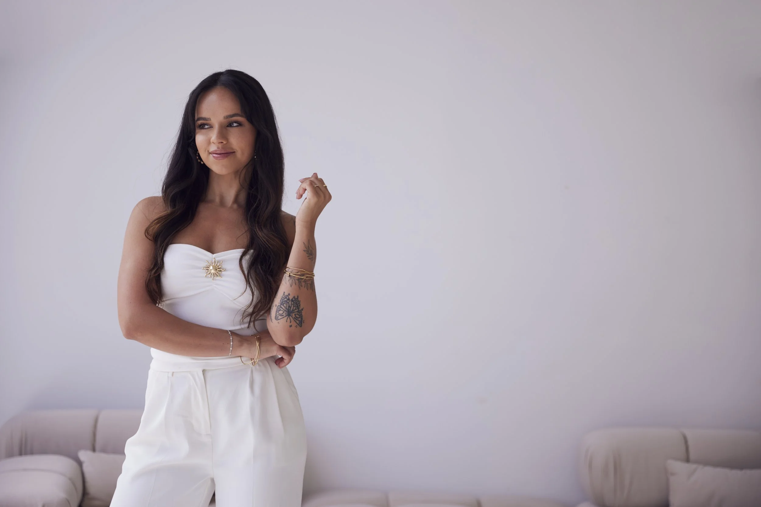 A woman with long dark hair and tattoos on her arm wearing a white top and white pants, standing in a room with a white wall and beige couch in the background.