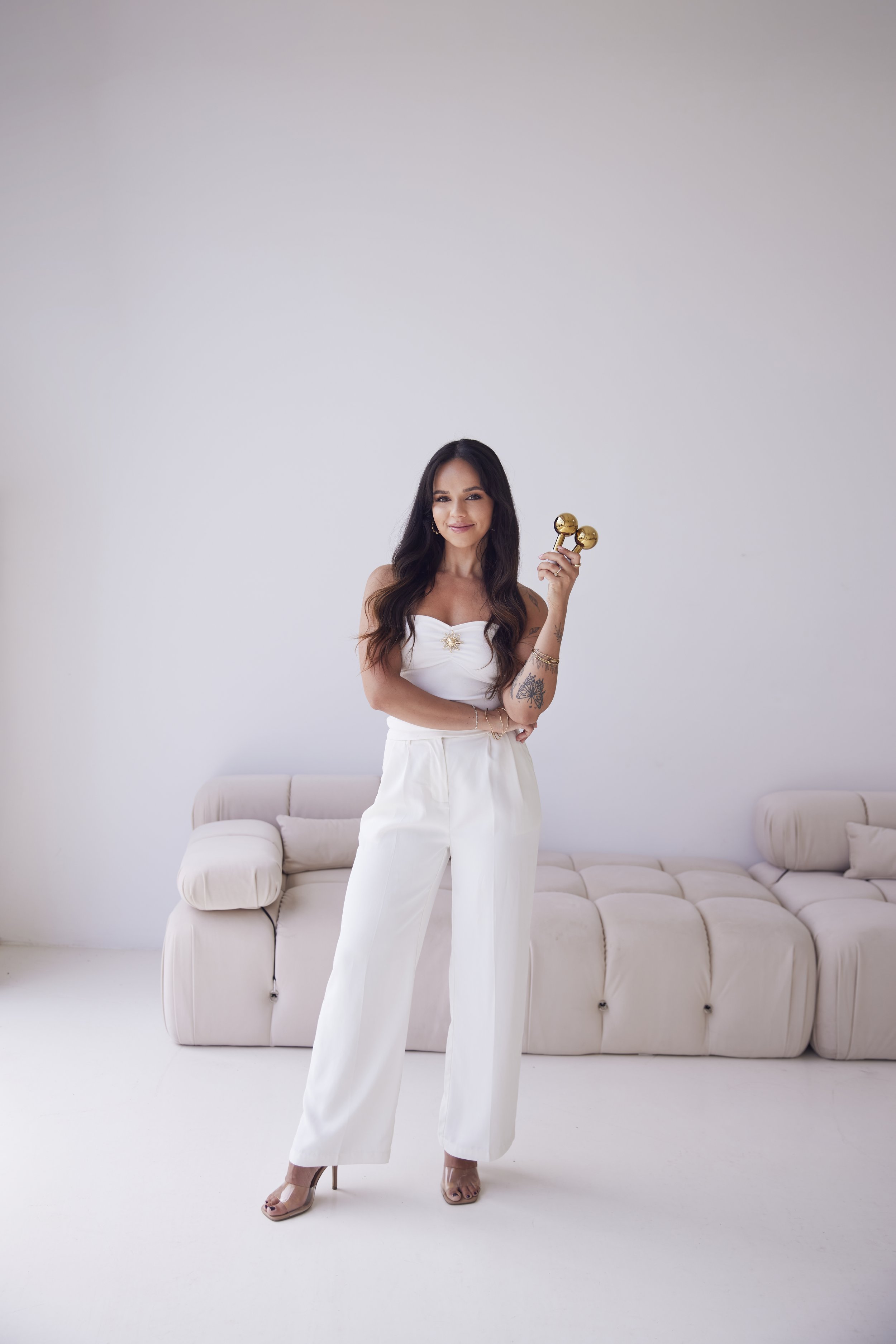 A woman with long dark hair, wearing a white strapless top and white wide-leg pants, is standing in a minimalist room. She is smiling and holding a golden massage roller in her right hand. The background features a white wall and a beige curved sofa.