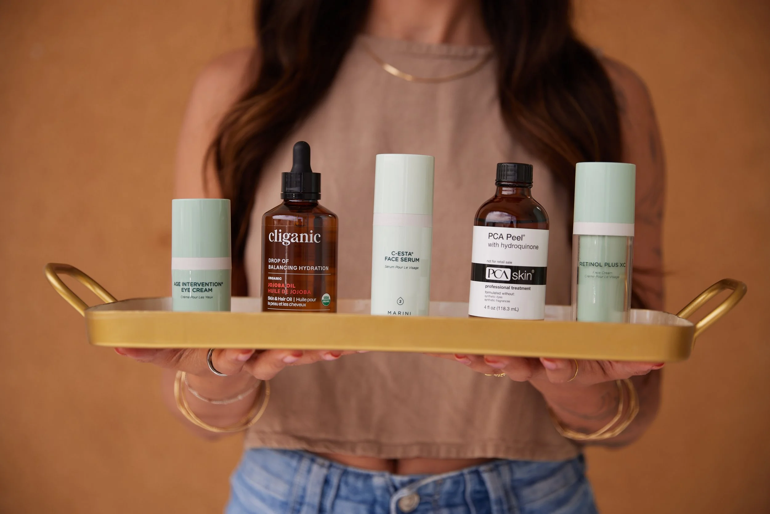 Person holding a gold tray with various skincare products against a brown background.