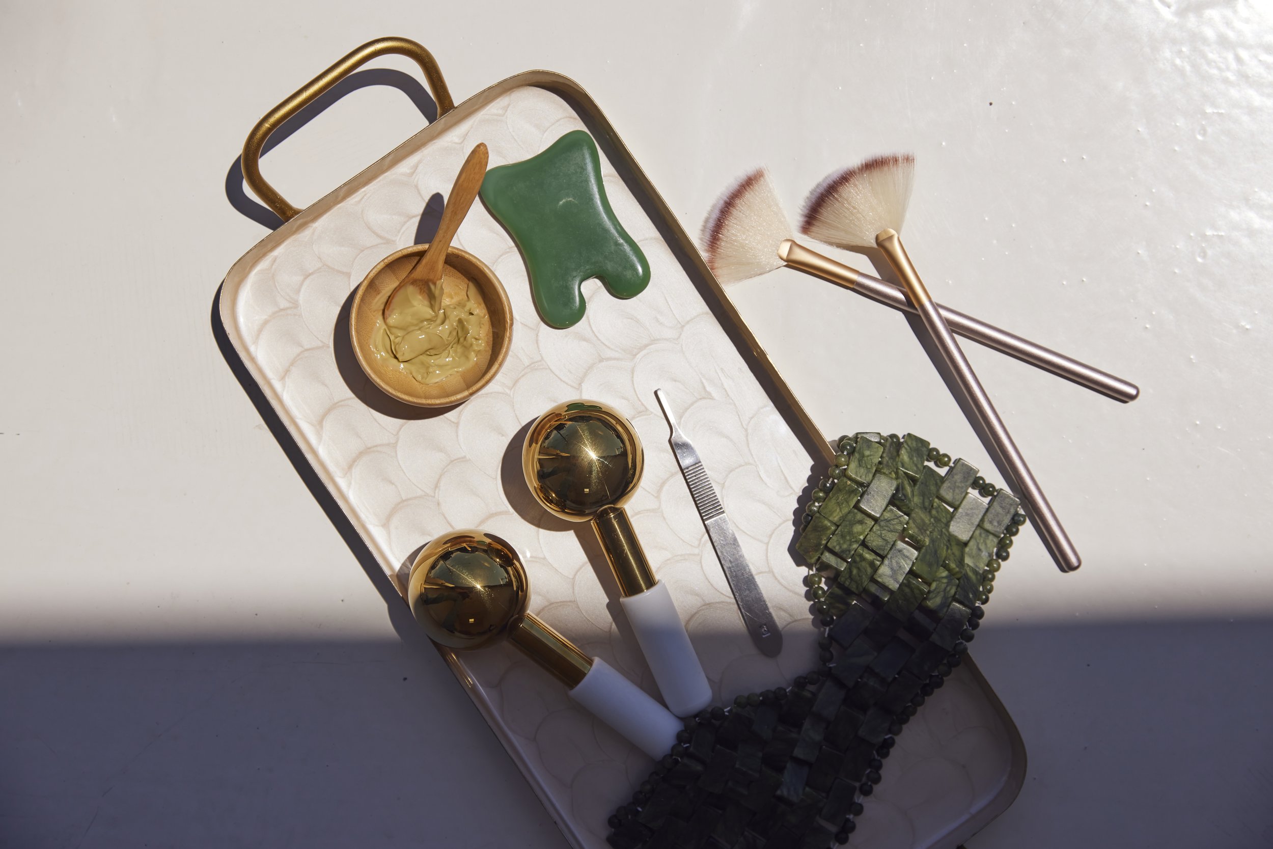 A collection of beauty and jewelry tools including a golden mirror, a pair of face brushes with light-colored bristles, a small bowl of cream or foundation with a small wooden spatula, a green shape resembling a dog, and a green stone bracelet, all arranged on a rectangular tray with a textured white surface.