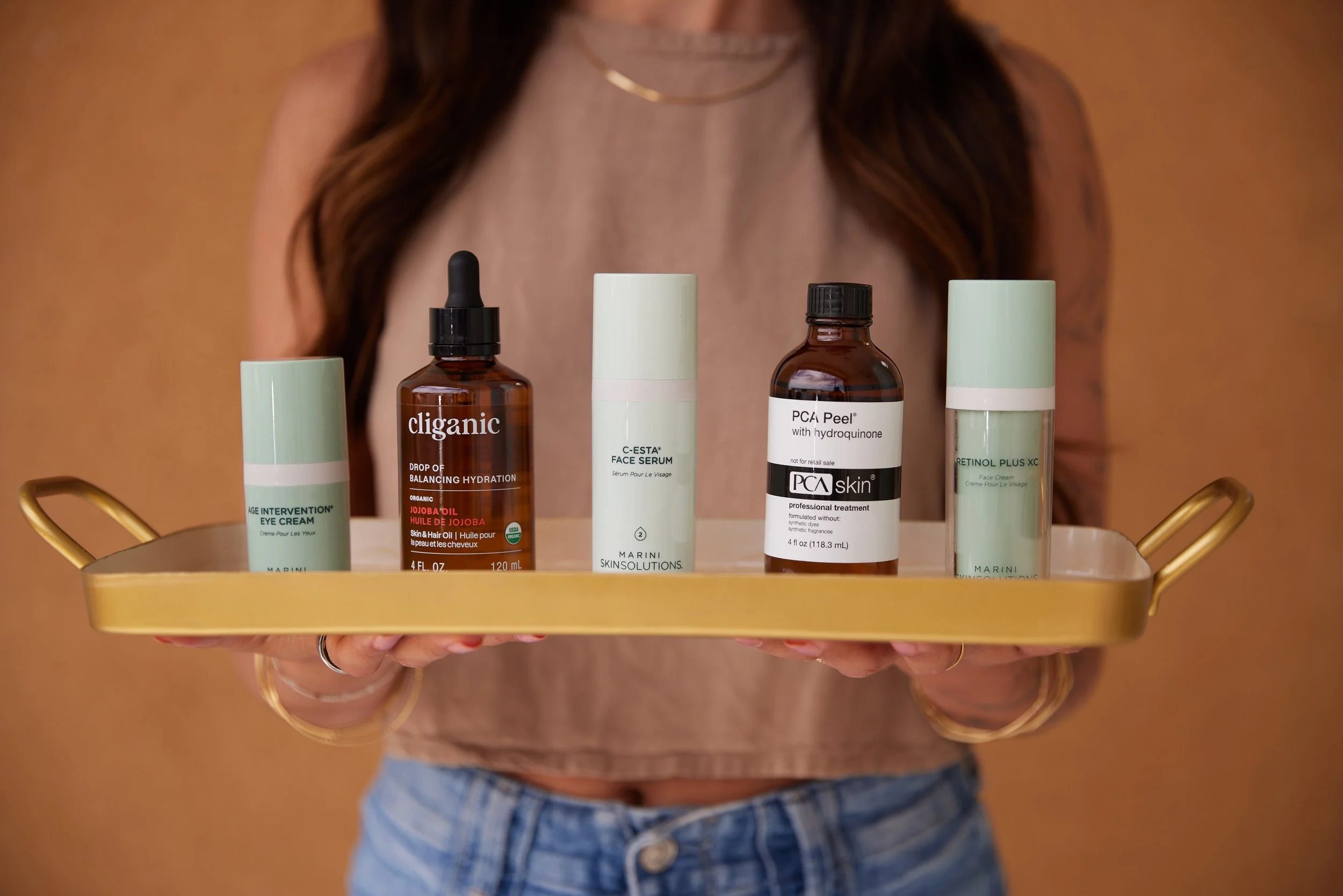 A person wearing a pink top and blue jeans holding a gold tray with skincare products, including eye cream, face serum, and facial treatment bottles, against a brown background.