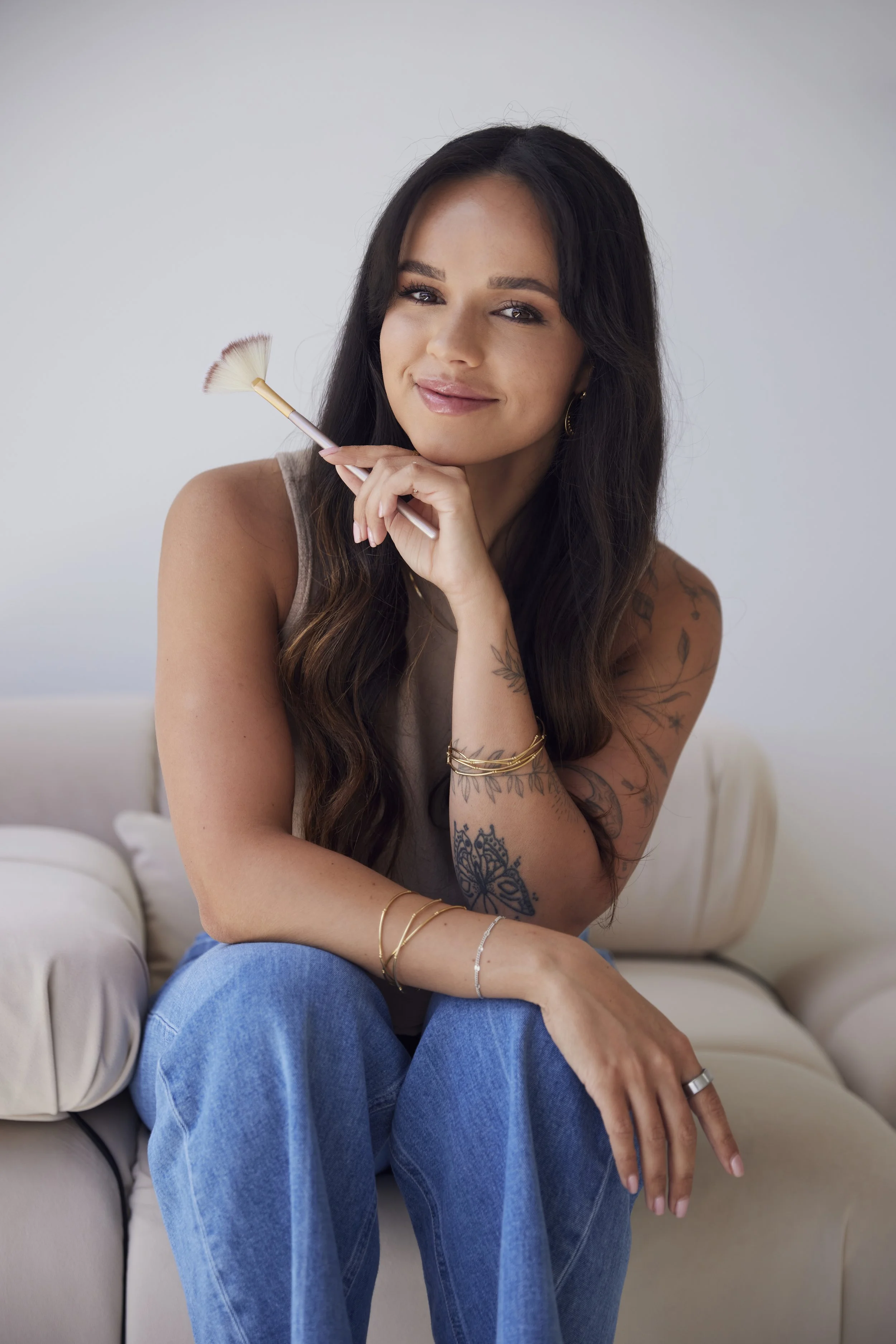 A woman with long dark hair and tattoos on her arms sitting on a beige couch, holding a makeup brush near her face and smiling at the camera.