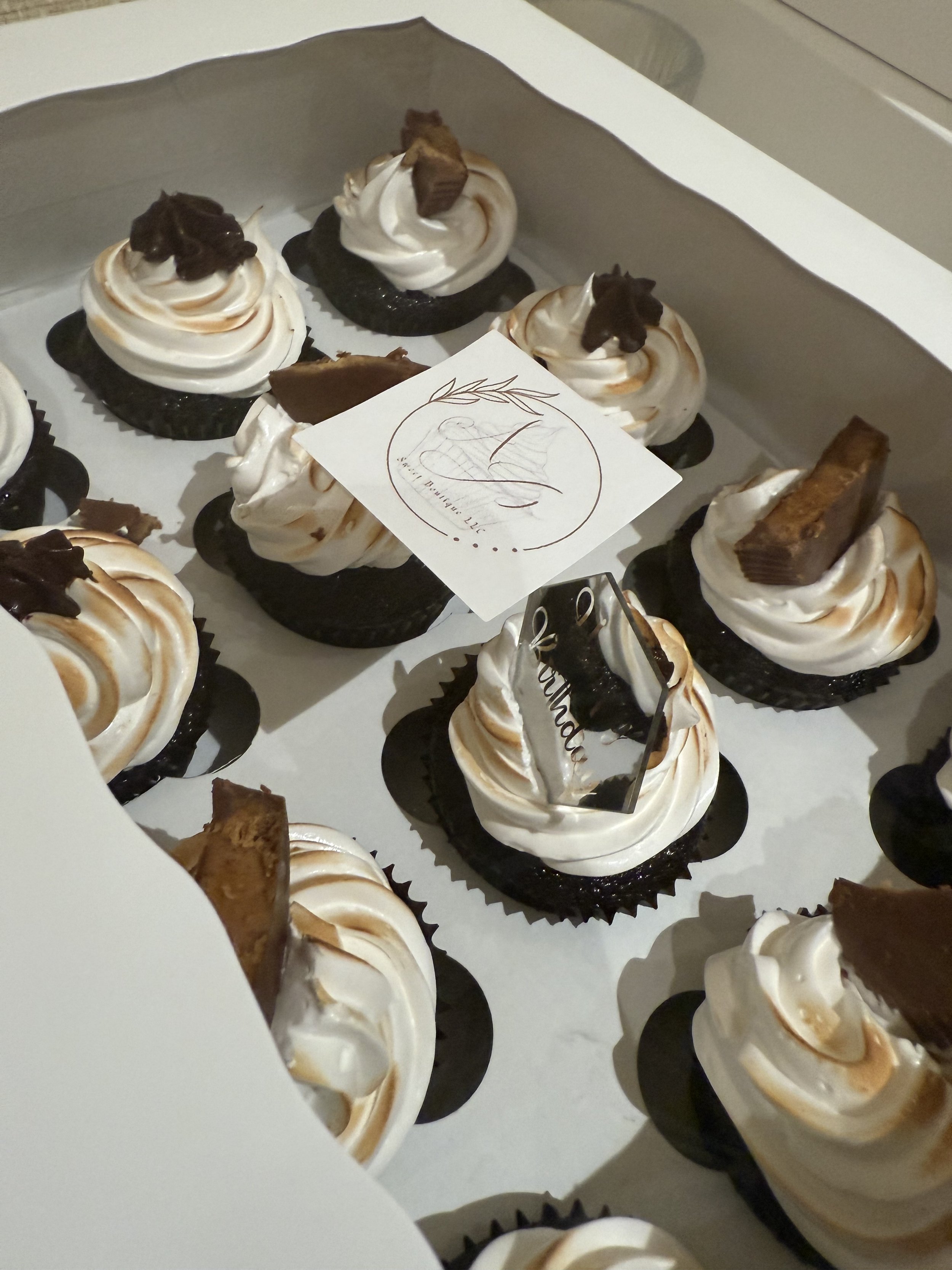 Box of chocolate cupcakes with swirled vanilla frosting, chocolate decorations, and a small decorative card in a white box.