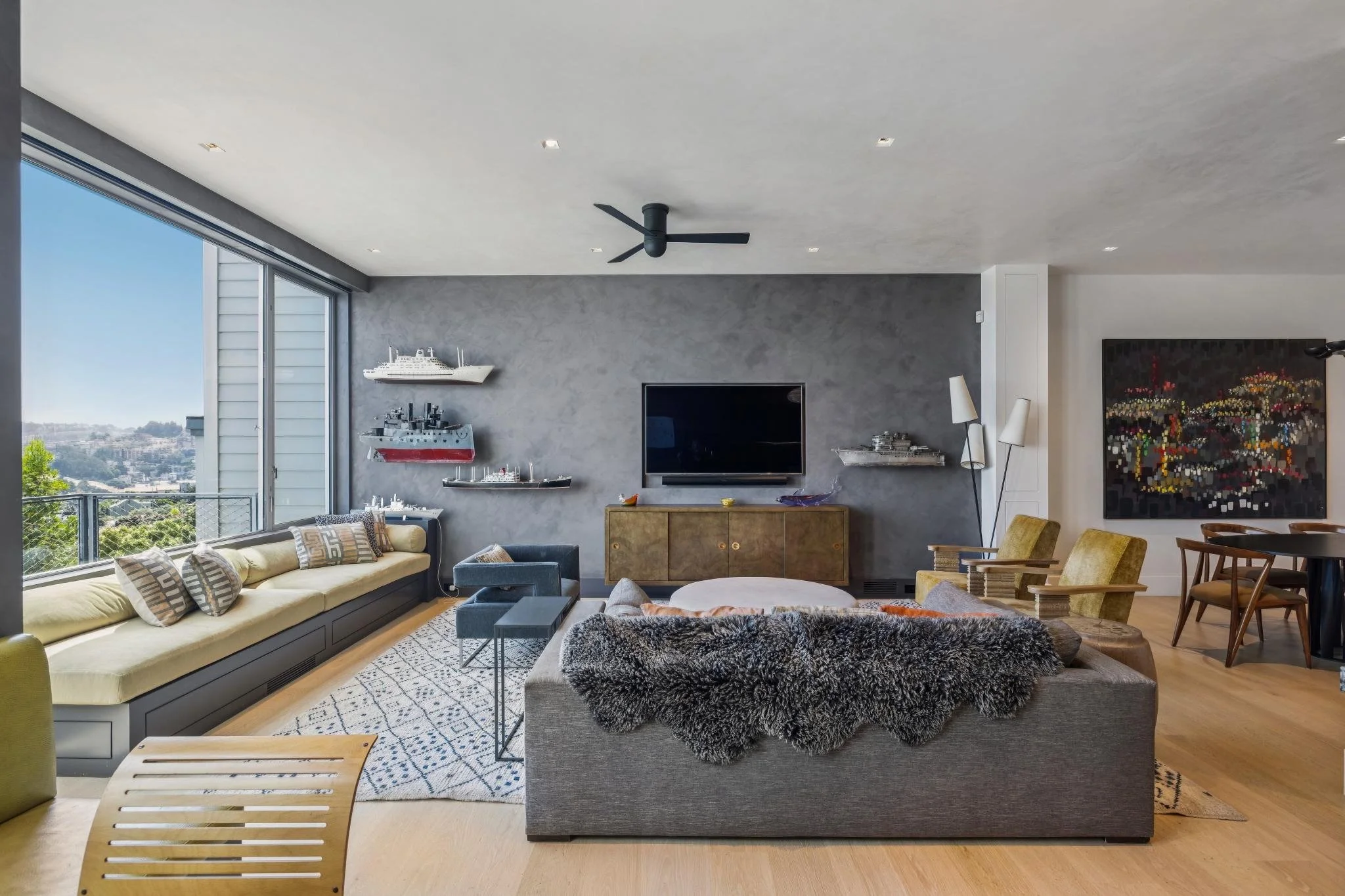 Spacious living room with large windows showing a city view, modern furniture including a beige sofa with patterned pillows, a gray armchair with a faux fur blanket, a wooden sideboard with model ships, a large flat-screen TV, paintings on the walls,