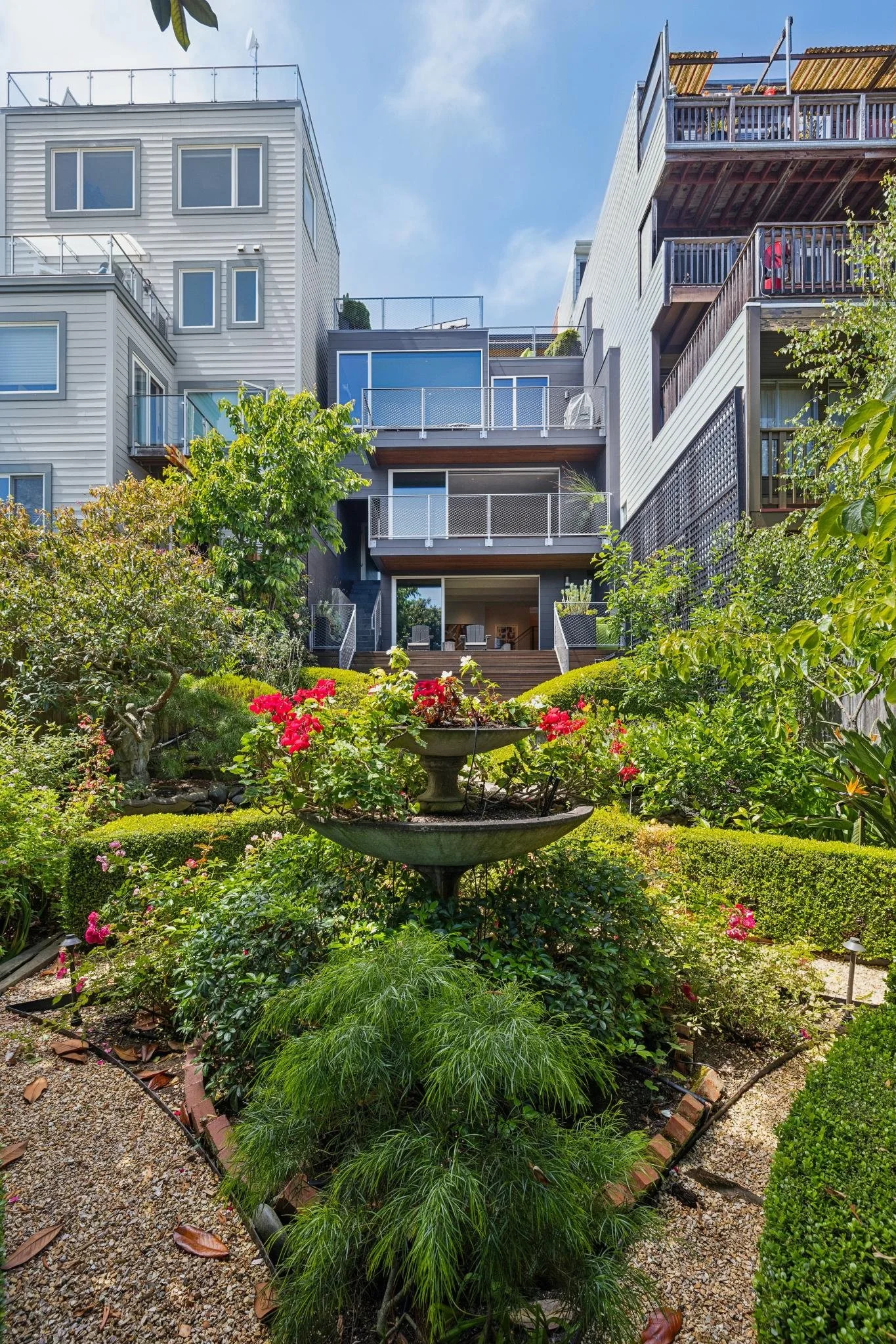 A multi-story residential apartment building with balconies, surrounded by a lush garden with various trees, bushes, flowers, and a decorative tiered fountain in the center.