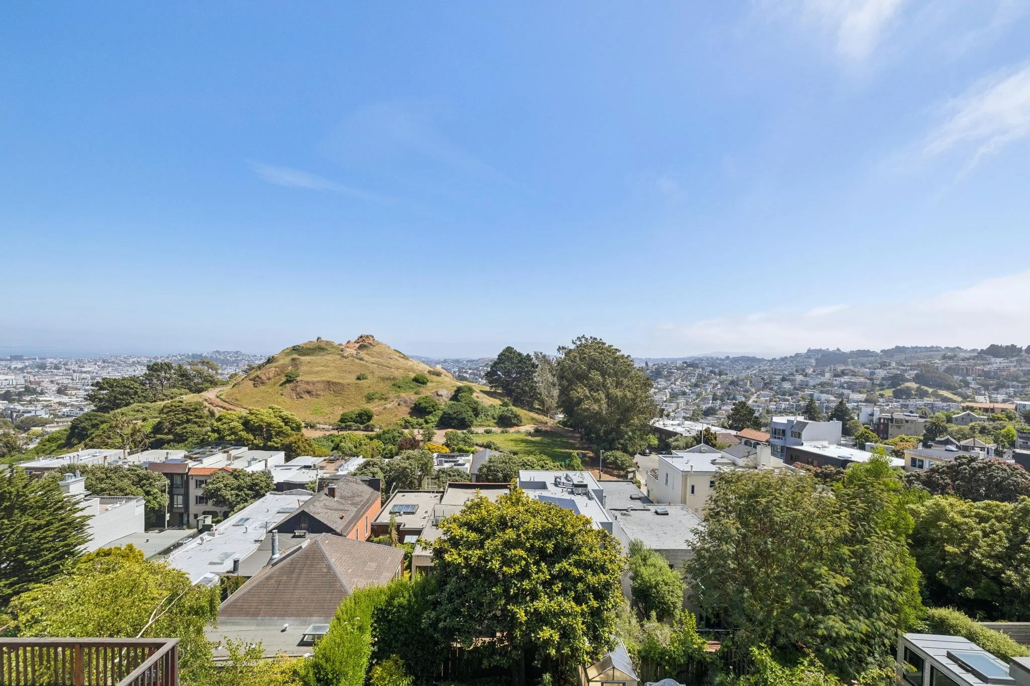 View of a hilly park with grass and trees, cityscape with residential buildings, and a bright blue sky with some clouds in the background.
