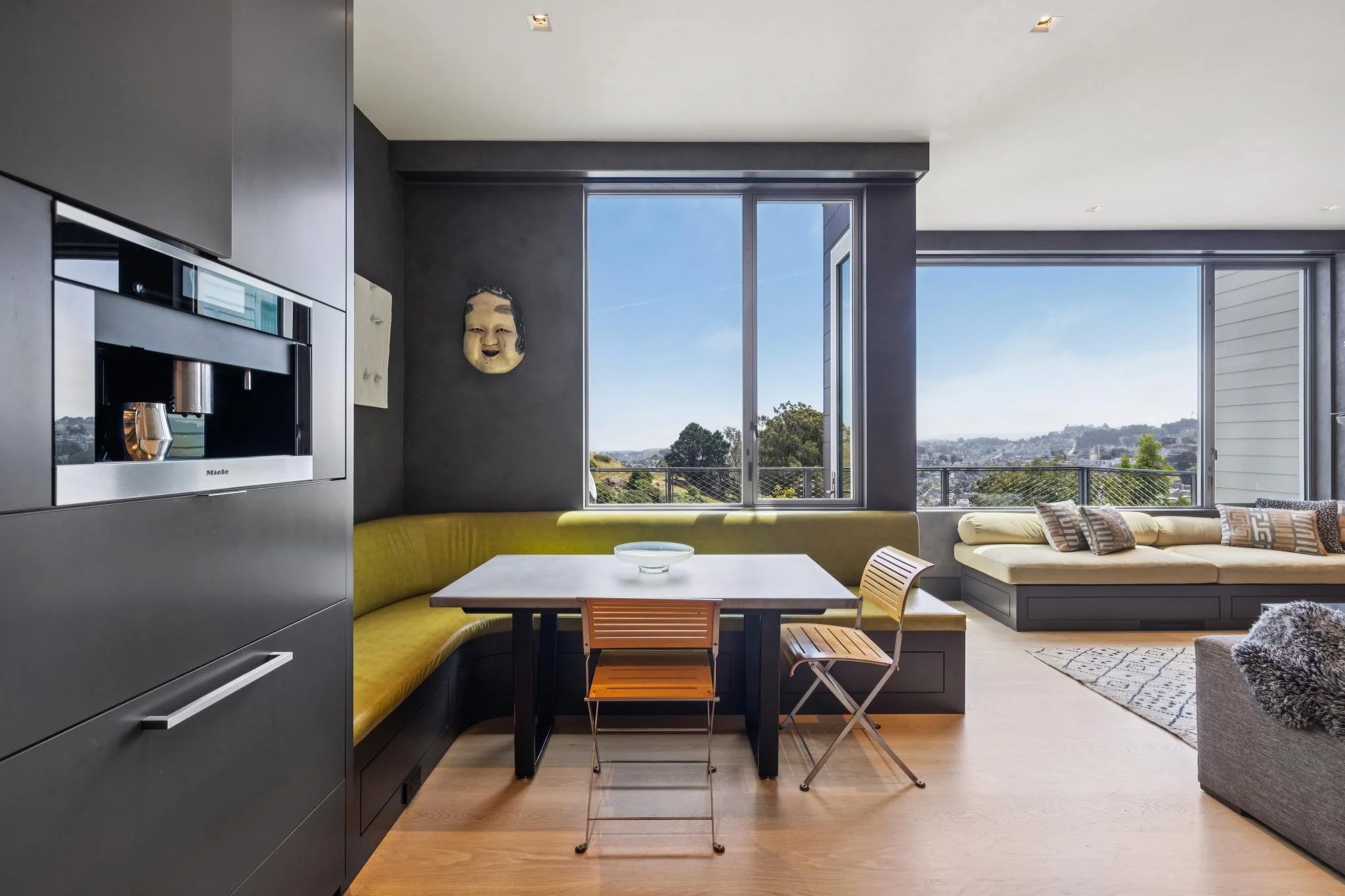 Interior view of a modern living room with large windows, yellow curved sofa, wooden table, chairs, and a textured gray sofa, with cityscape visible outside.
