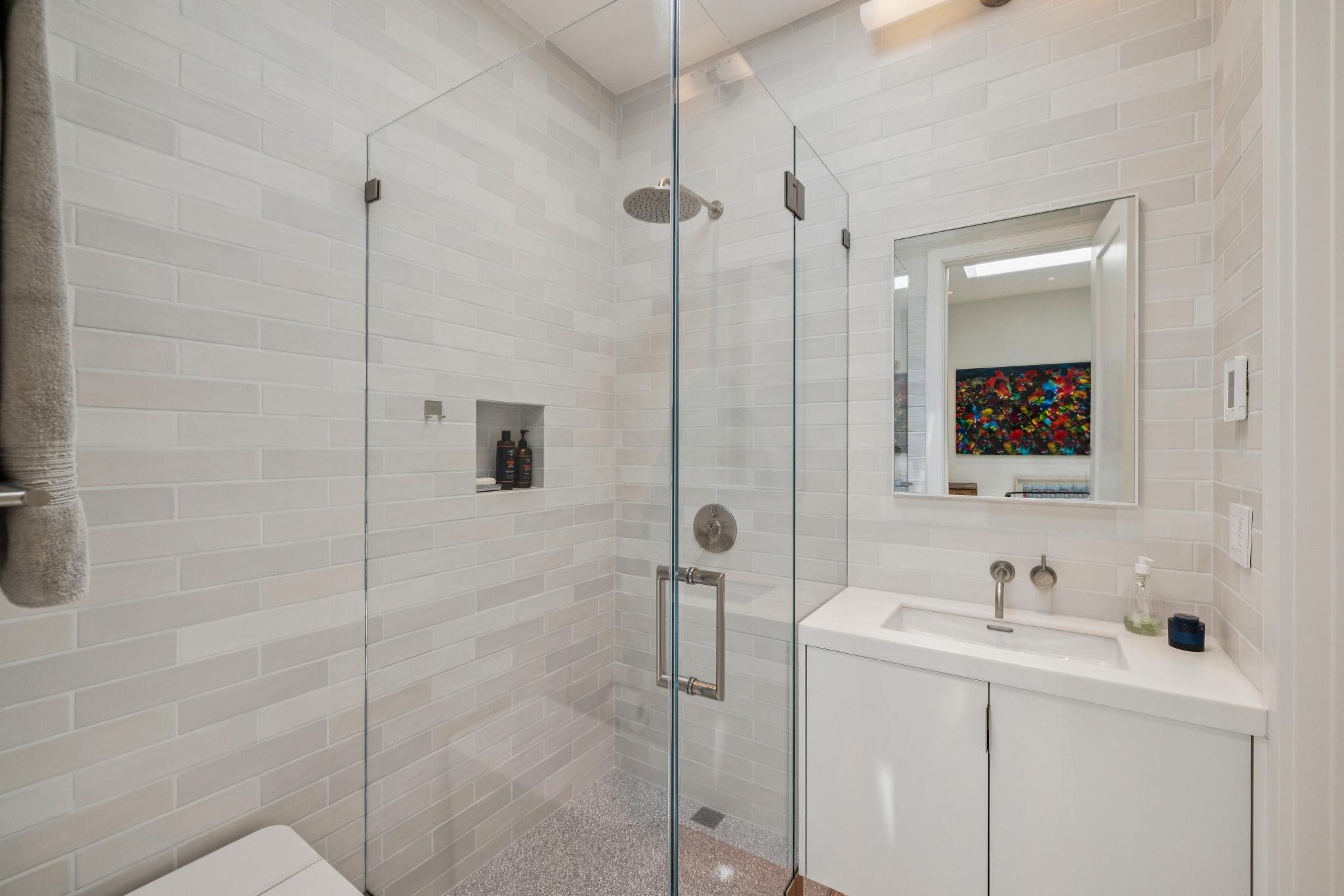 A modern bathroom with a walk-in shower enclosed by glass, white brick-patterned tiles on the walls, a showerhead, and a built-in shelf with toiletries. A mirror above the sink reflects artwork on the opposite wall.