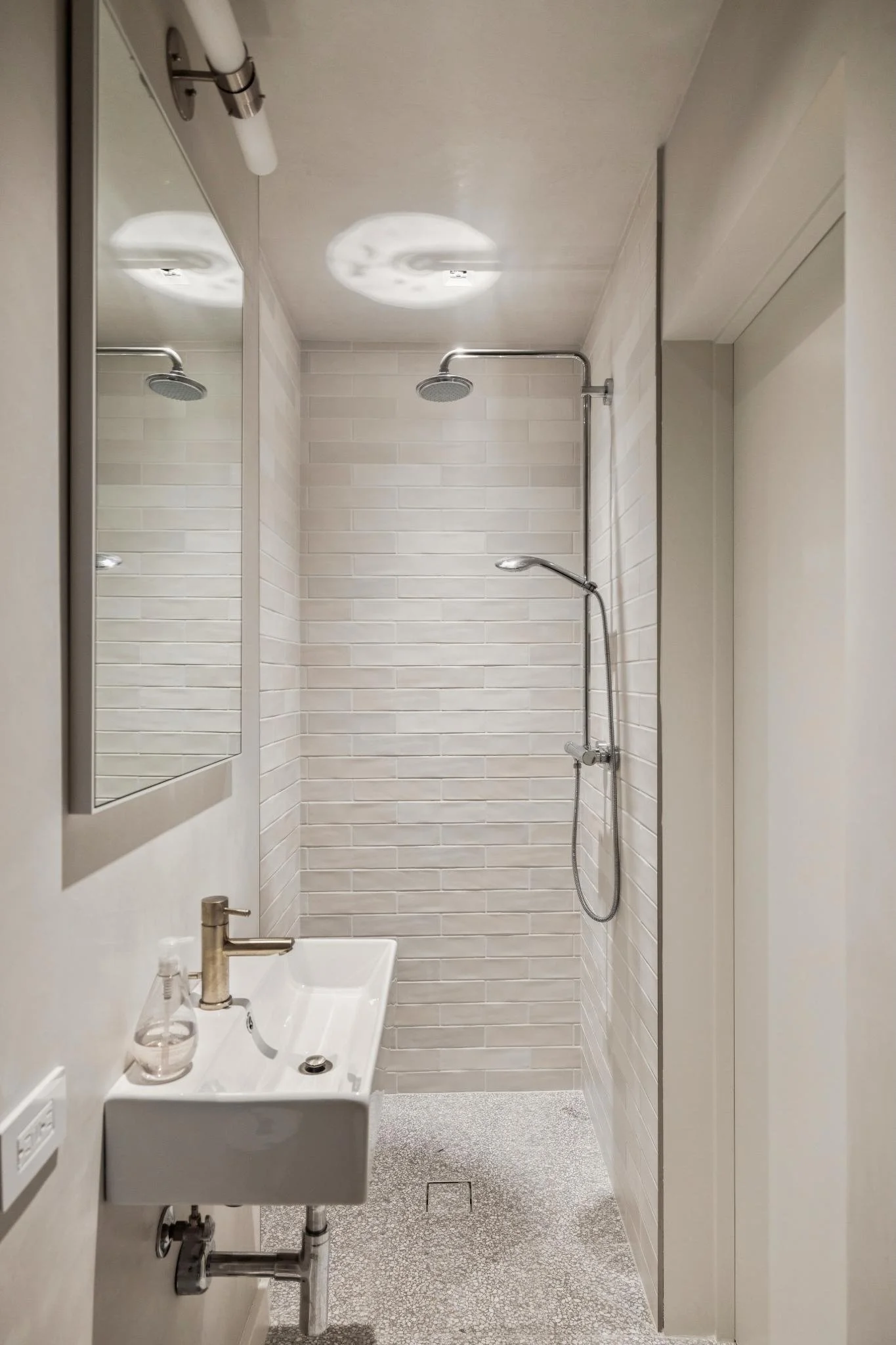 Small bathroom with white brick shower wall, ceiling shower head, and handheld shower wand, white sink with soap dispenser, mirror, and minimal decor.