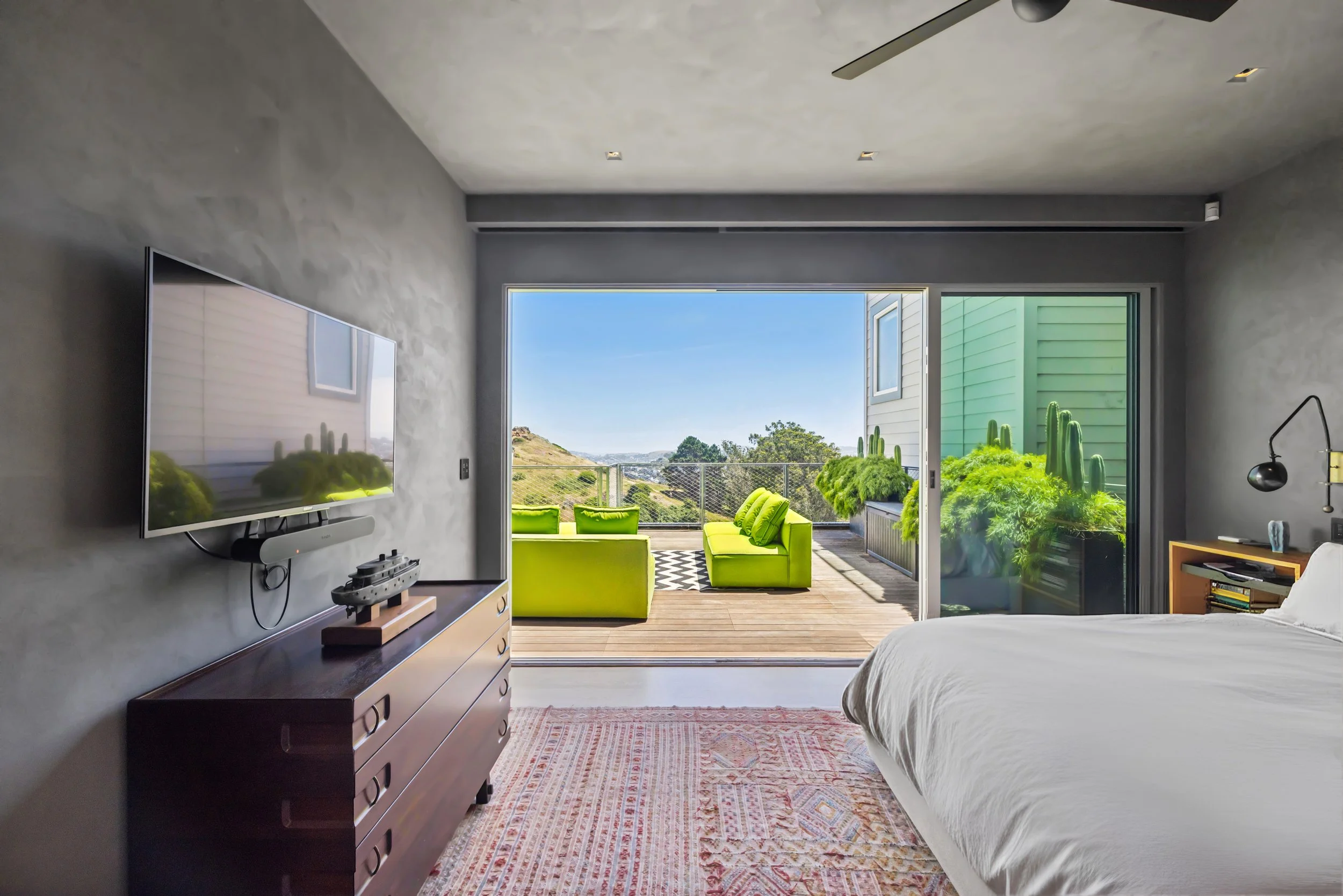 Interior of a bedroom with a large sliding glass door opening to a balcony with green outdoor furniture and a view of trees and hills in the distance. The room has a bed with white bedding, a wooden dresser, a mounted flat-screen TV, and a patterned 
