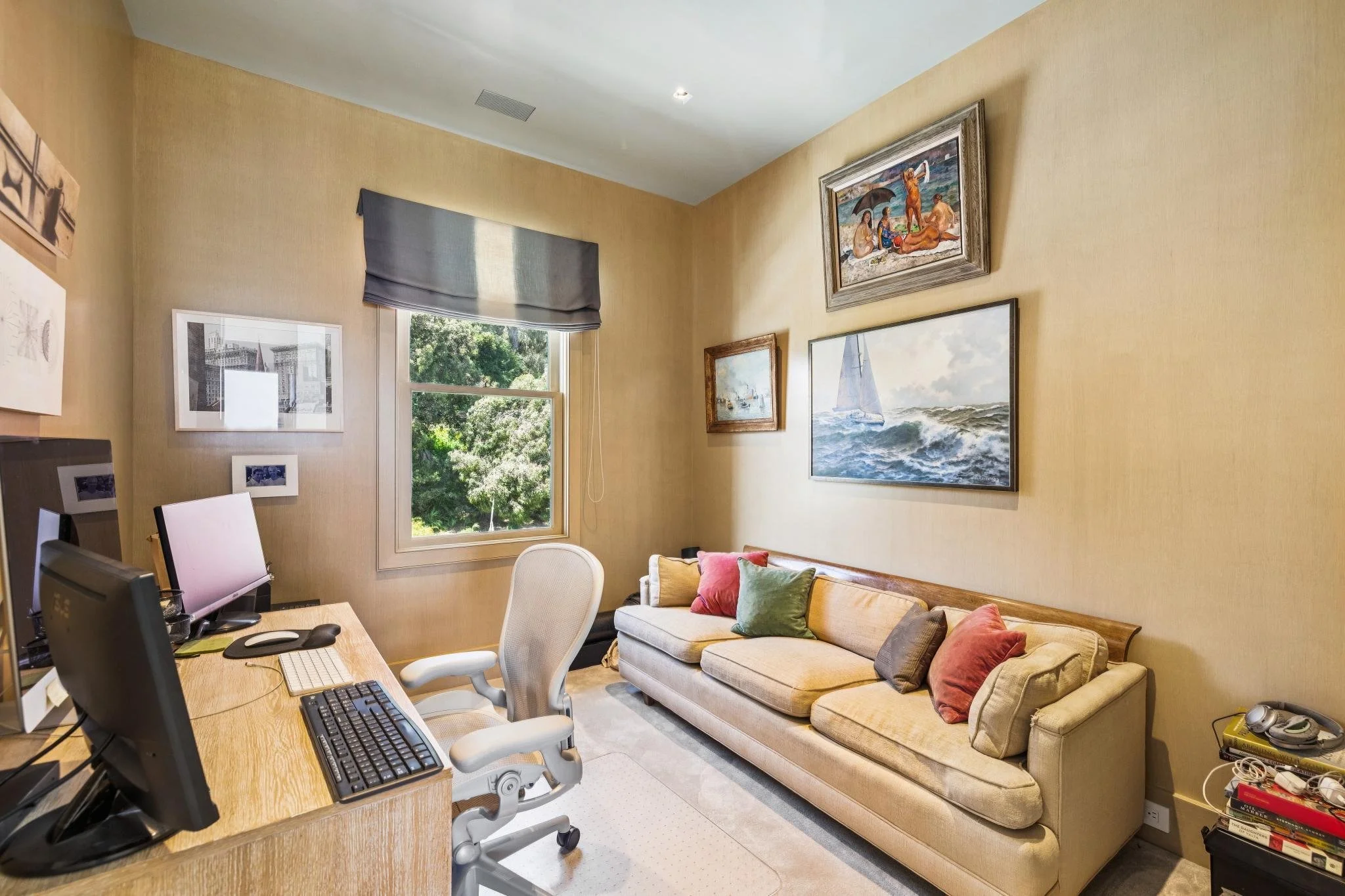A cozy office with a wooden desk, multiple computer monitors, and a white ergonomic chair. There's a beige sofa with colorful pillows and framed artwork on the walls, including ocean scenes and beach scenes with people. A window shows green trees out