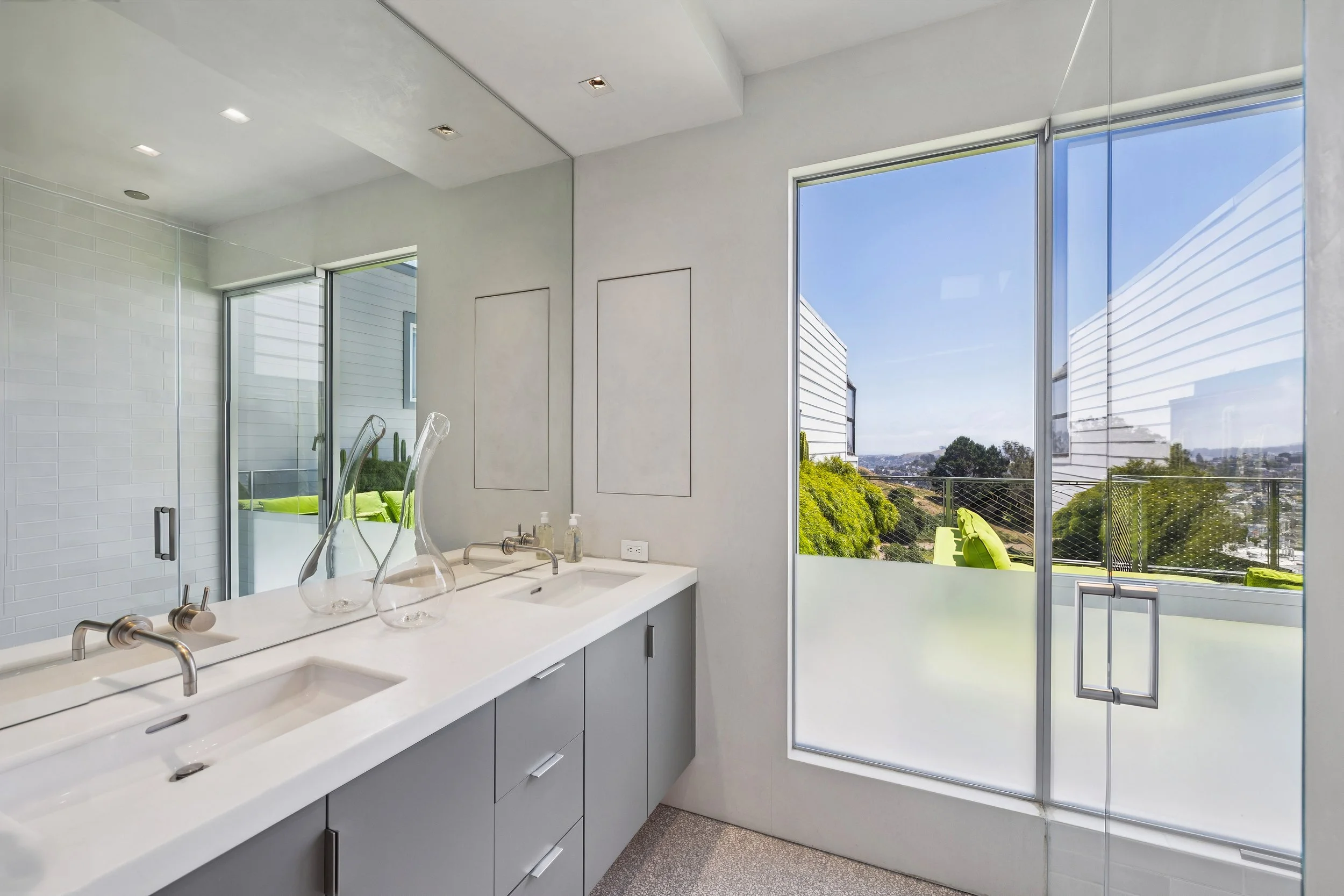 Modern bathroom with large windows, white vanity with sink, glass decor bottles, and outdoor view of greenery and houses.