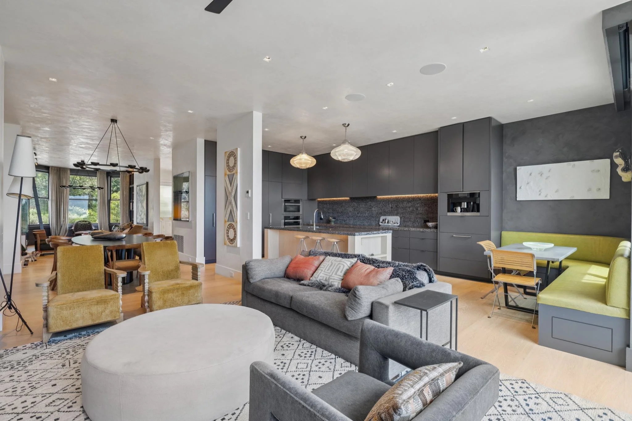 Interior view of a modern open-concept living space with a kitchen, dining area, and seating area, featuring gray, yellow, and neutral-colored furniture, pendant lights, and large windows.